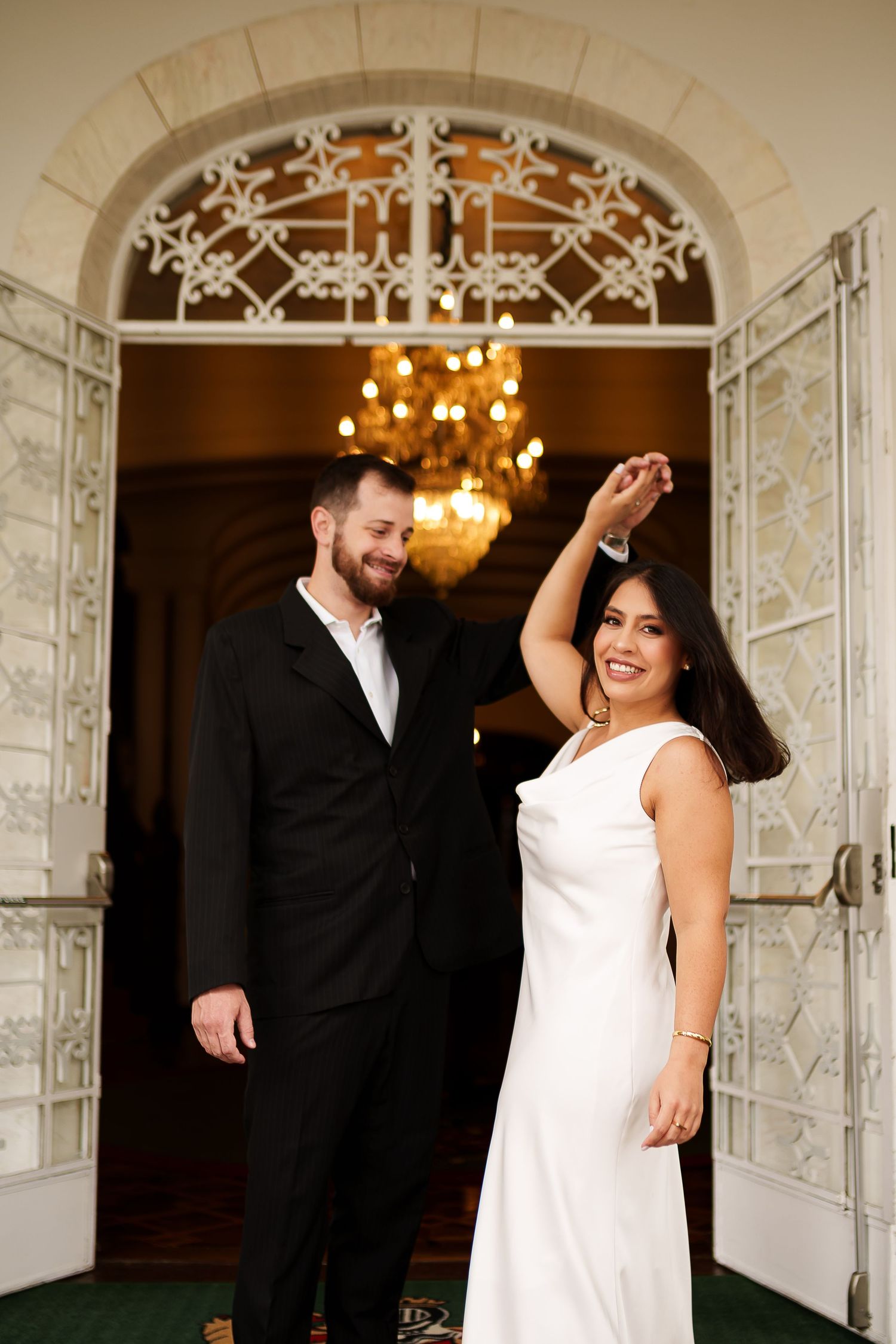 Couple in wedding attire dancing in front of ornate arched doors with chandelier, bride in white dress spinning joyfully.