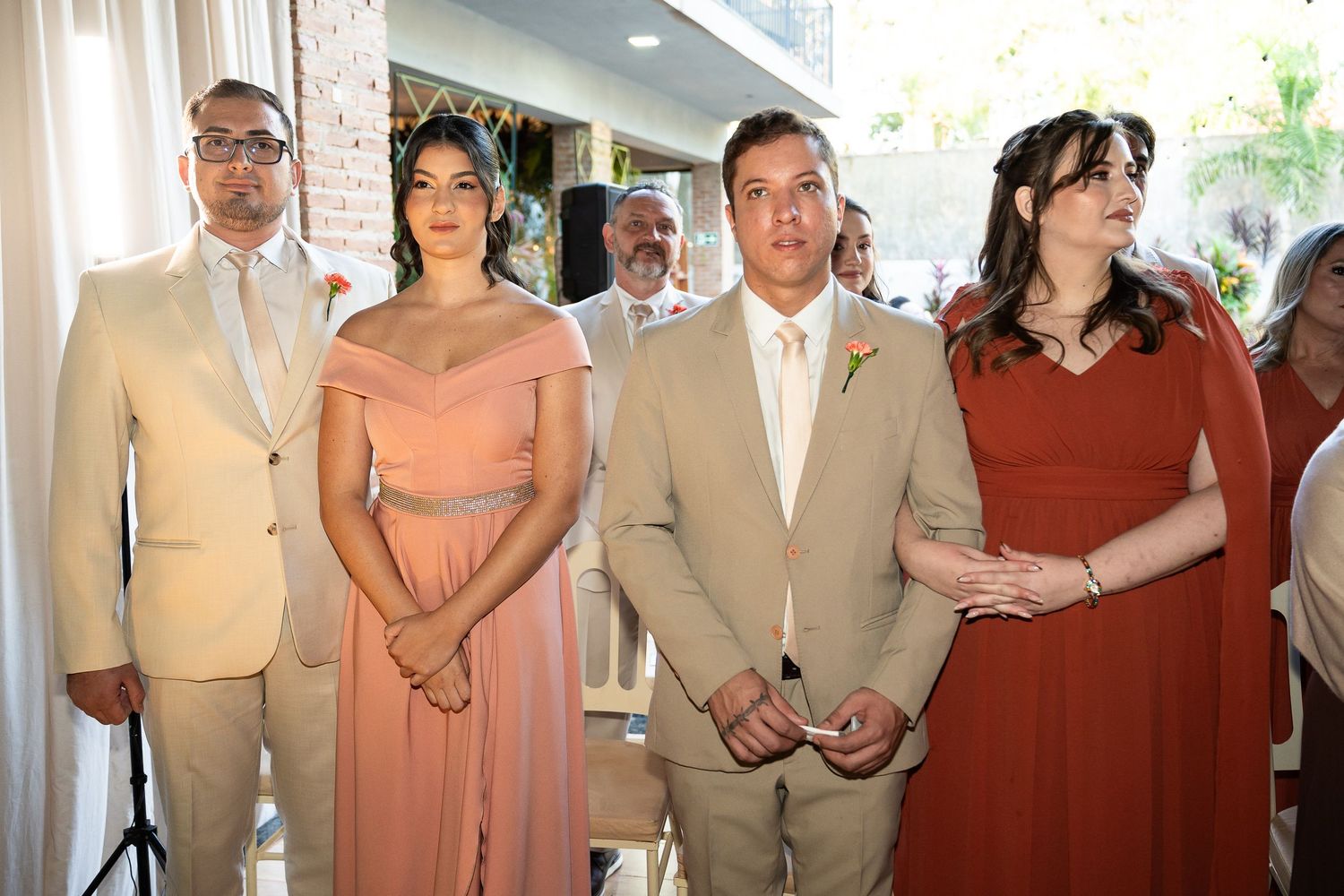 Two couples in formal wedding attire, men in tan suits, women in pink and red dresses, standing at a wedding ceremony.