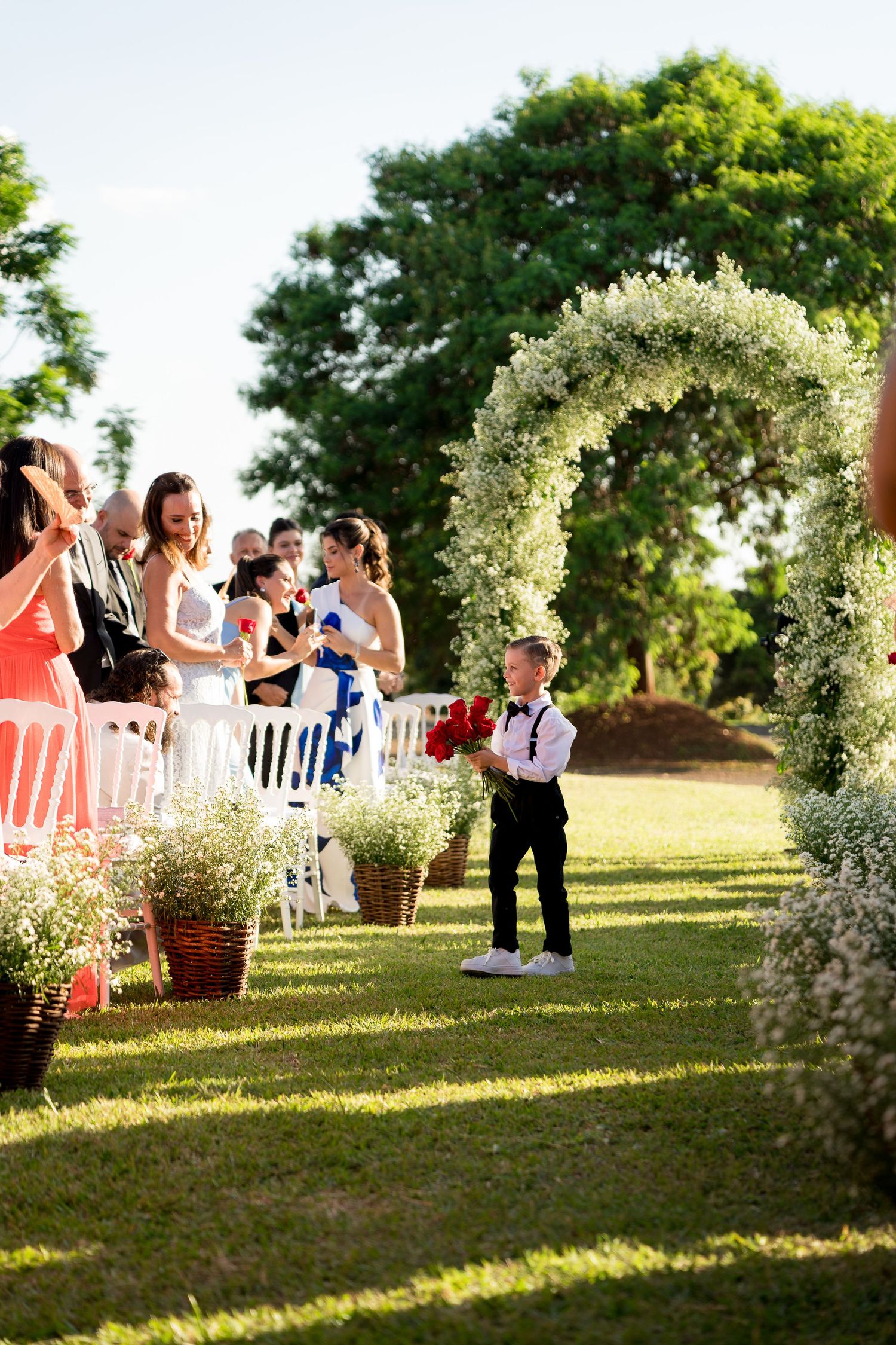 Young ring bearer in black suit carries red roses down floral arch aisle at outdoor wedding ceremony.