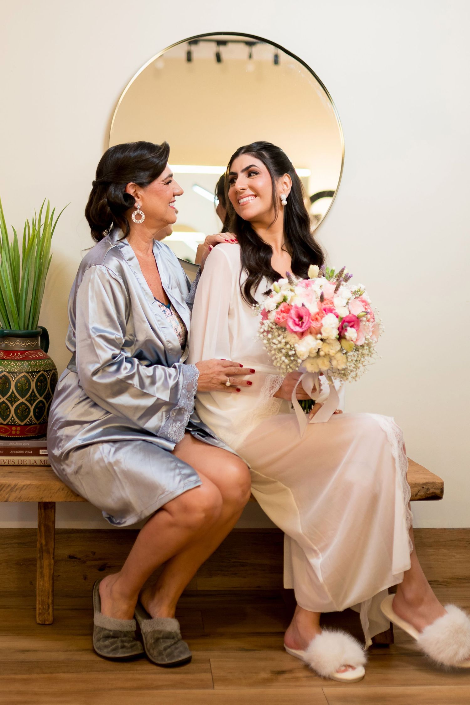Bride in satin robe holds pink bouquet sitting with older woman in silver robe on bench before mirror.