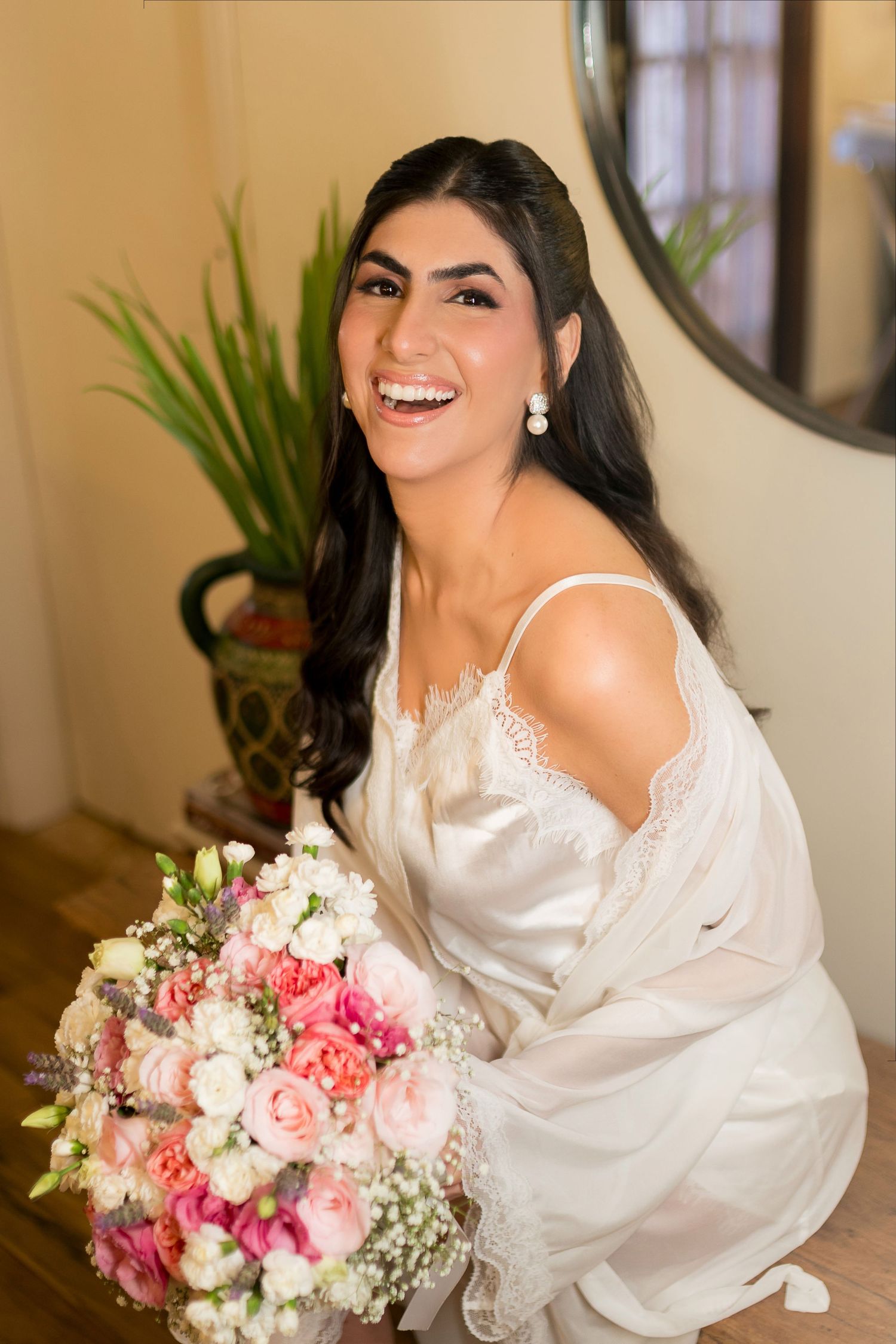 Smiling bride in white satin robe holding a pink and white floral bouquet, laughing joyfully during wedding preparations.