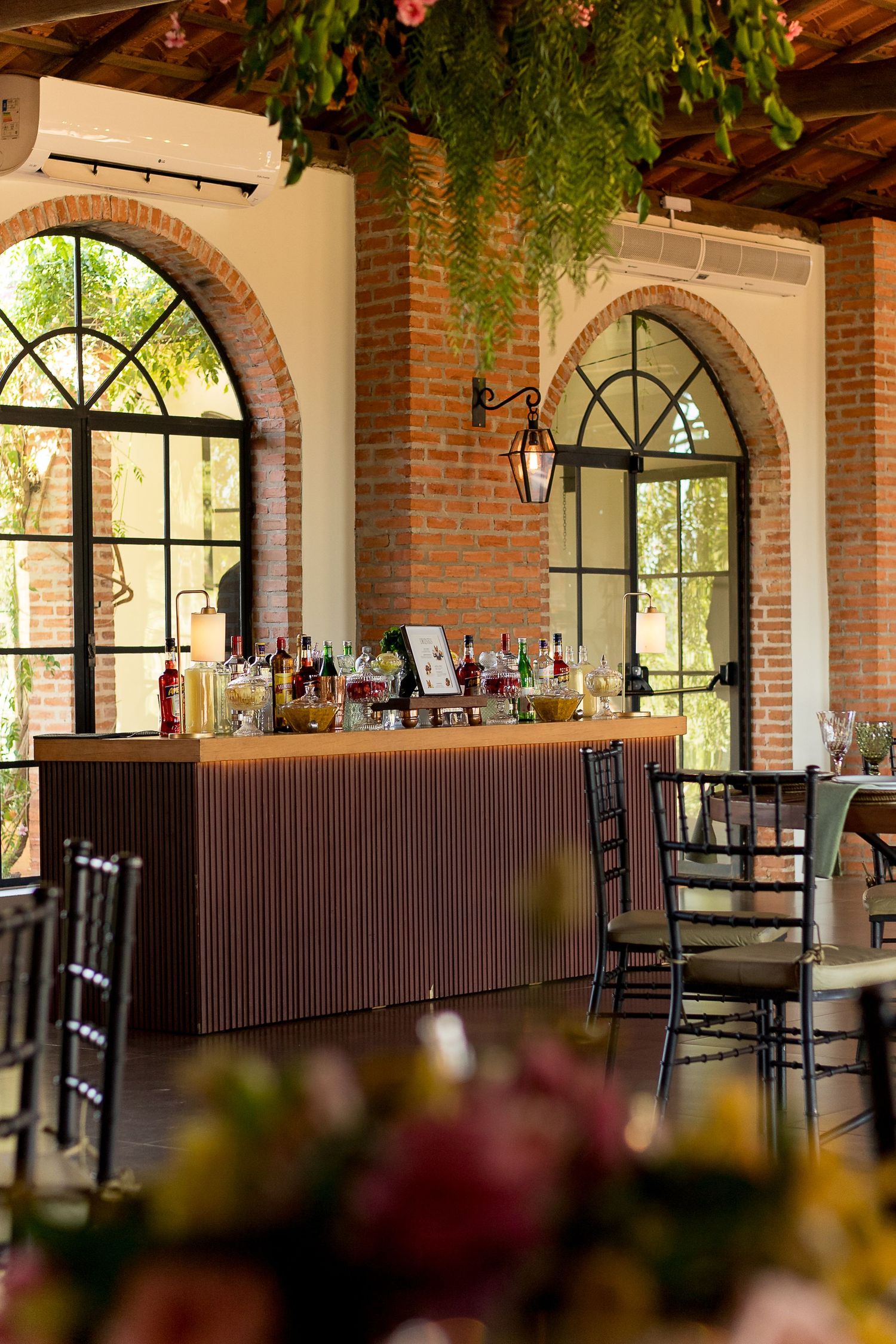 Elegant event venue with brick walls, arched windows, a decorated bar, black chairs, and lush greenery overhead.