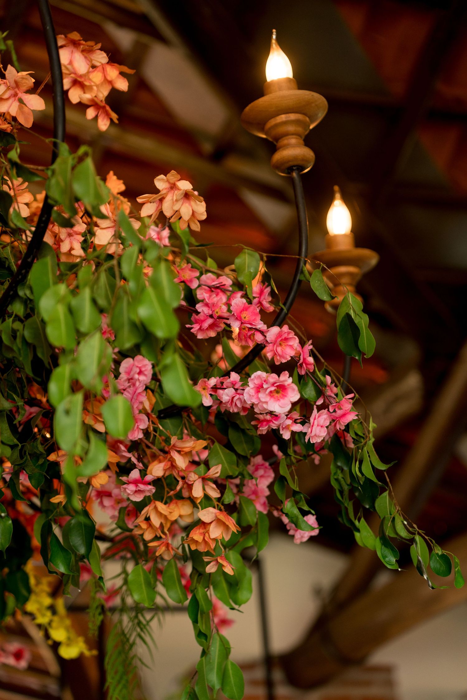 Pink and orange flowers cascade over a decorative black metal stand with glowing candle lights in a warmly lit rustic interior.
