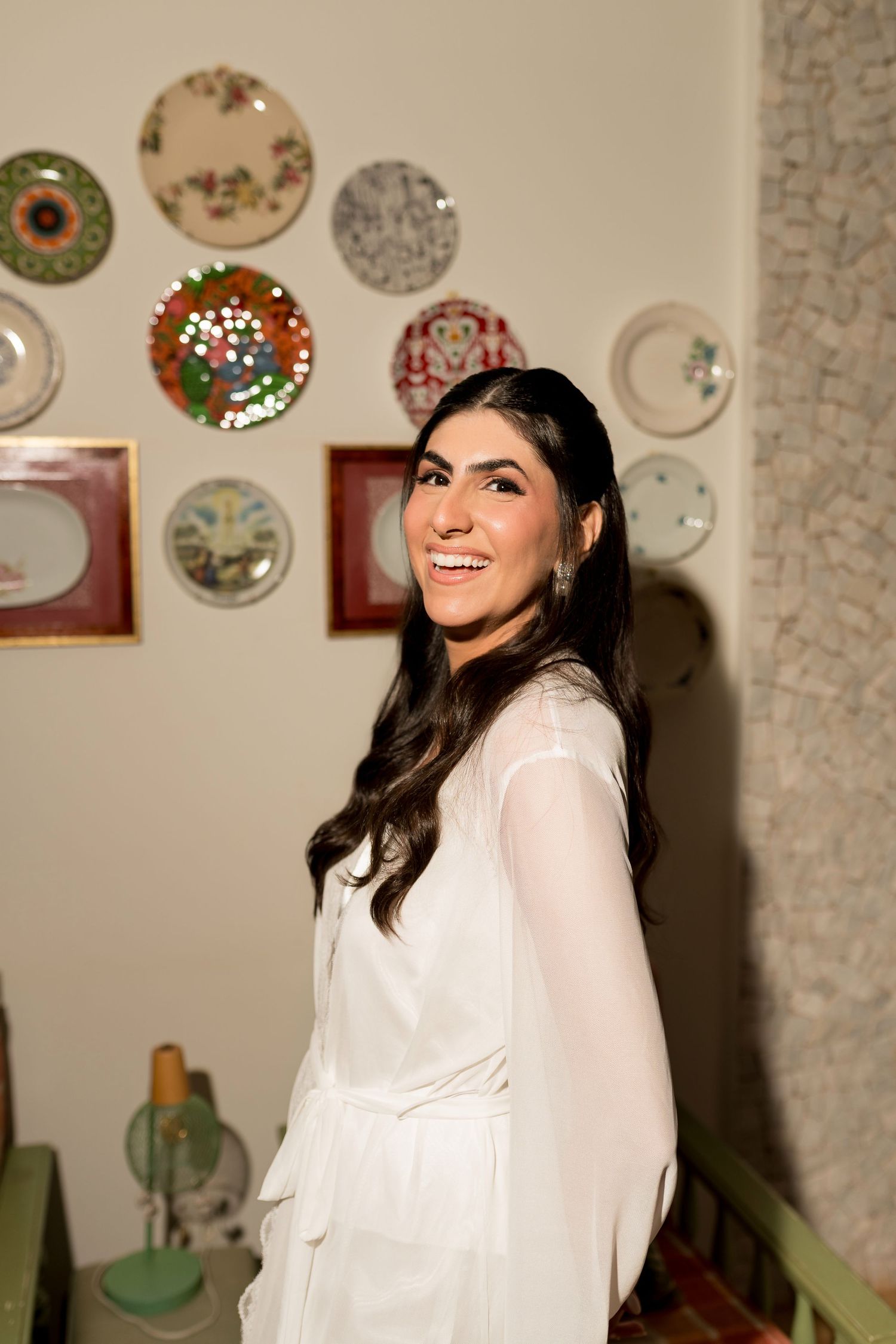 Smiling woman in white dress poses indoors with decorative plates displayed on wall behind her.