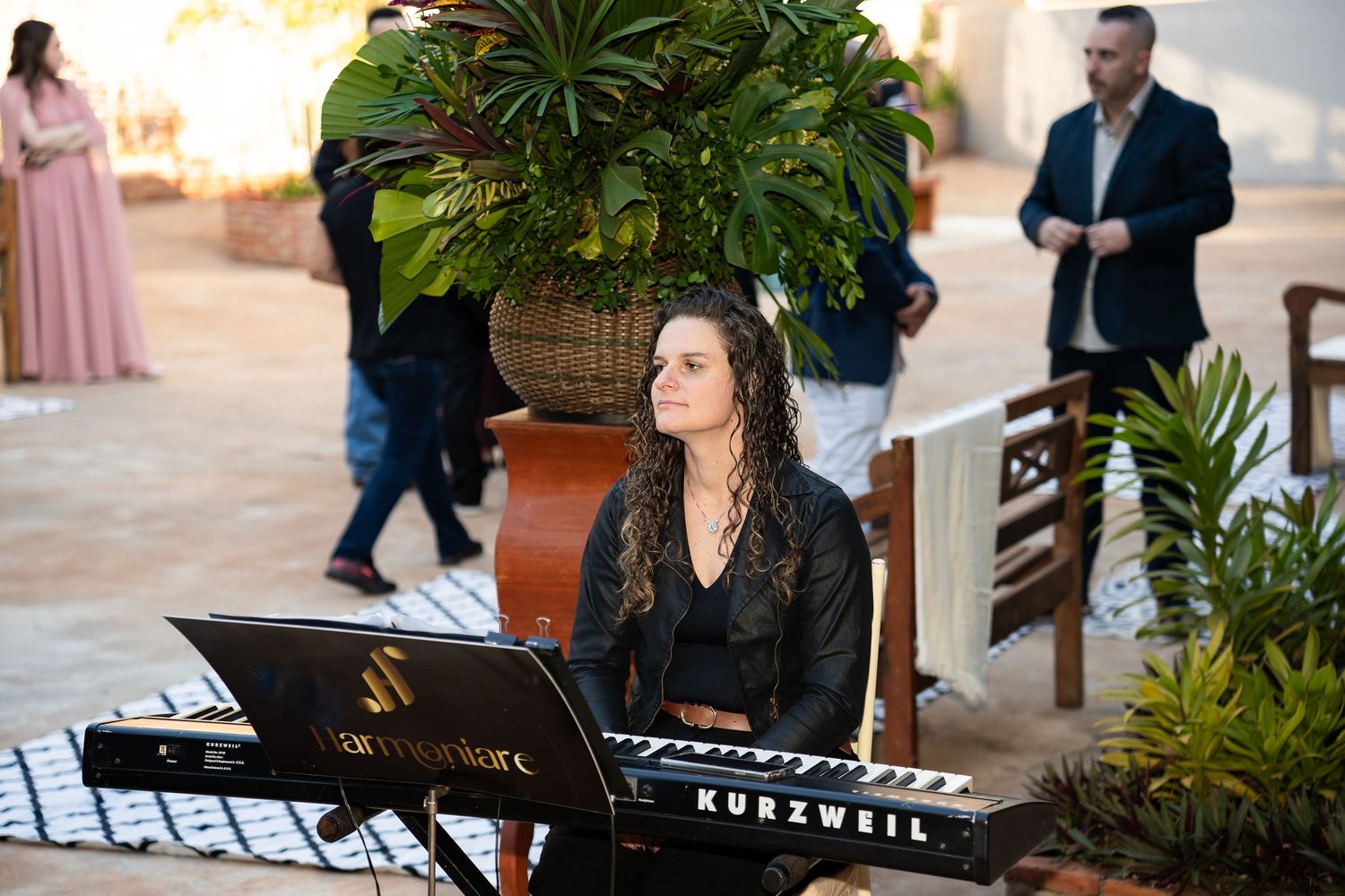Woman playing a Kurzweil keyboard at an outdoor wedding reception surrounded by tropical plants and guests.
