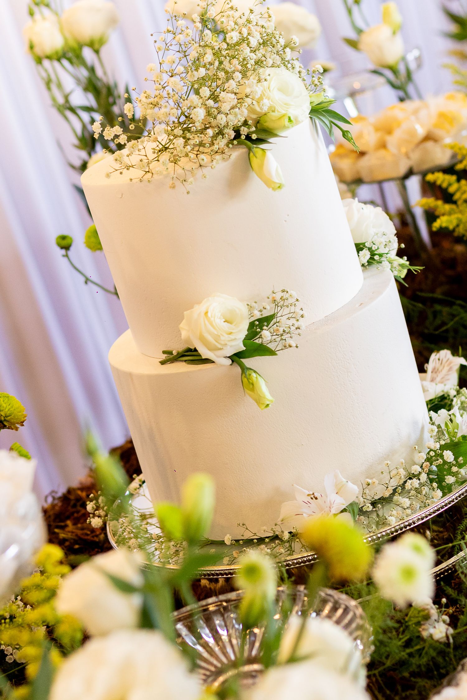 Elegant two-tier white wedding cake adorned with fresh white roses and baby's breath, surrounded by floral arrangements.