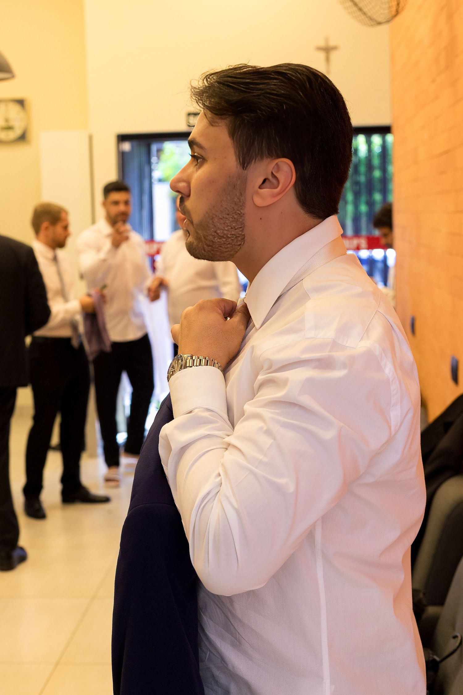 A well-dressed man in a white shirt adjusts his tie while others in formal attire stand in the background.