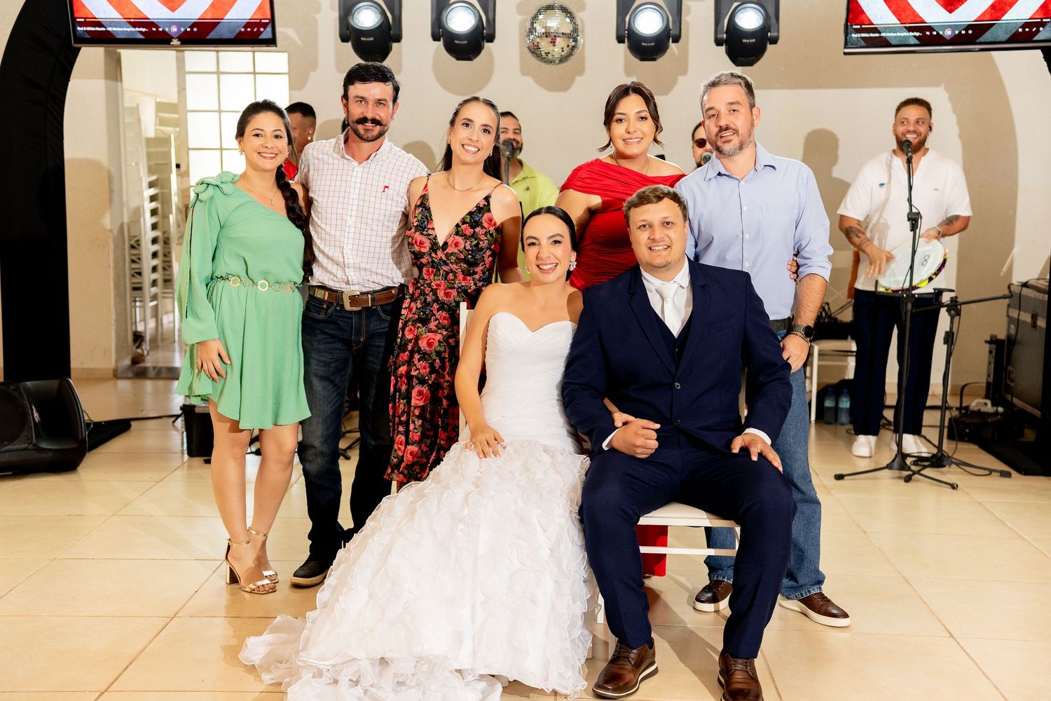 Wedding couple poses with guests in elegant venue, bride in white gown and groom in navy suit, surrounded by smiling friends.