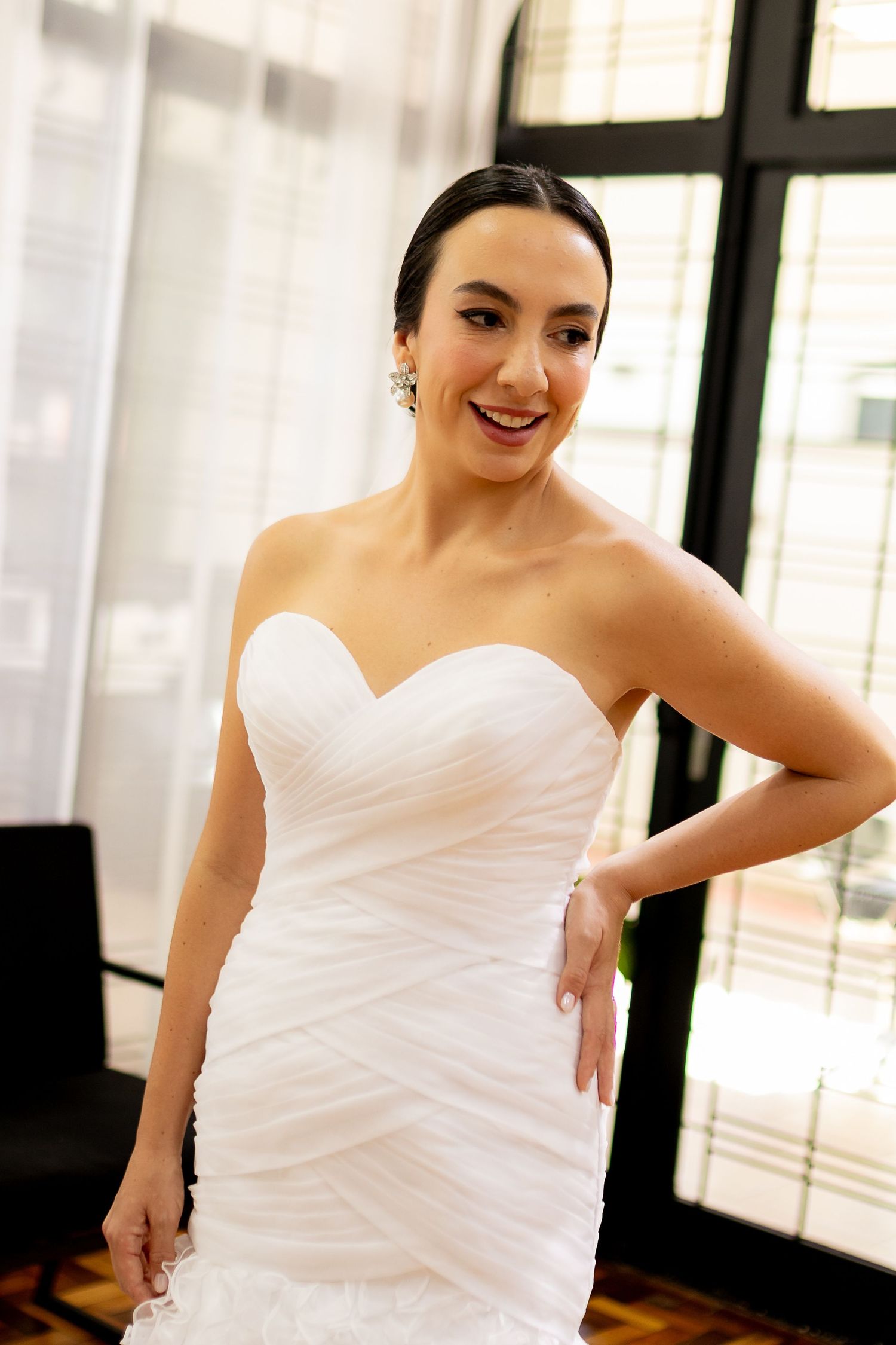 Smiling bride in a strapless white ruched sweetheart gown posing with hand on hip near a window.