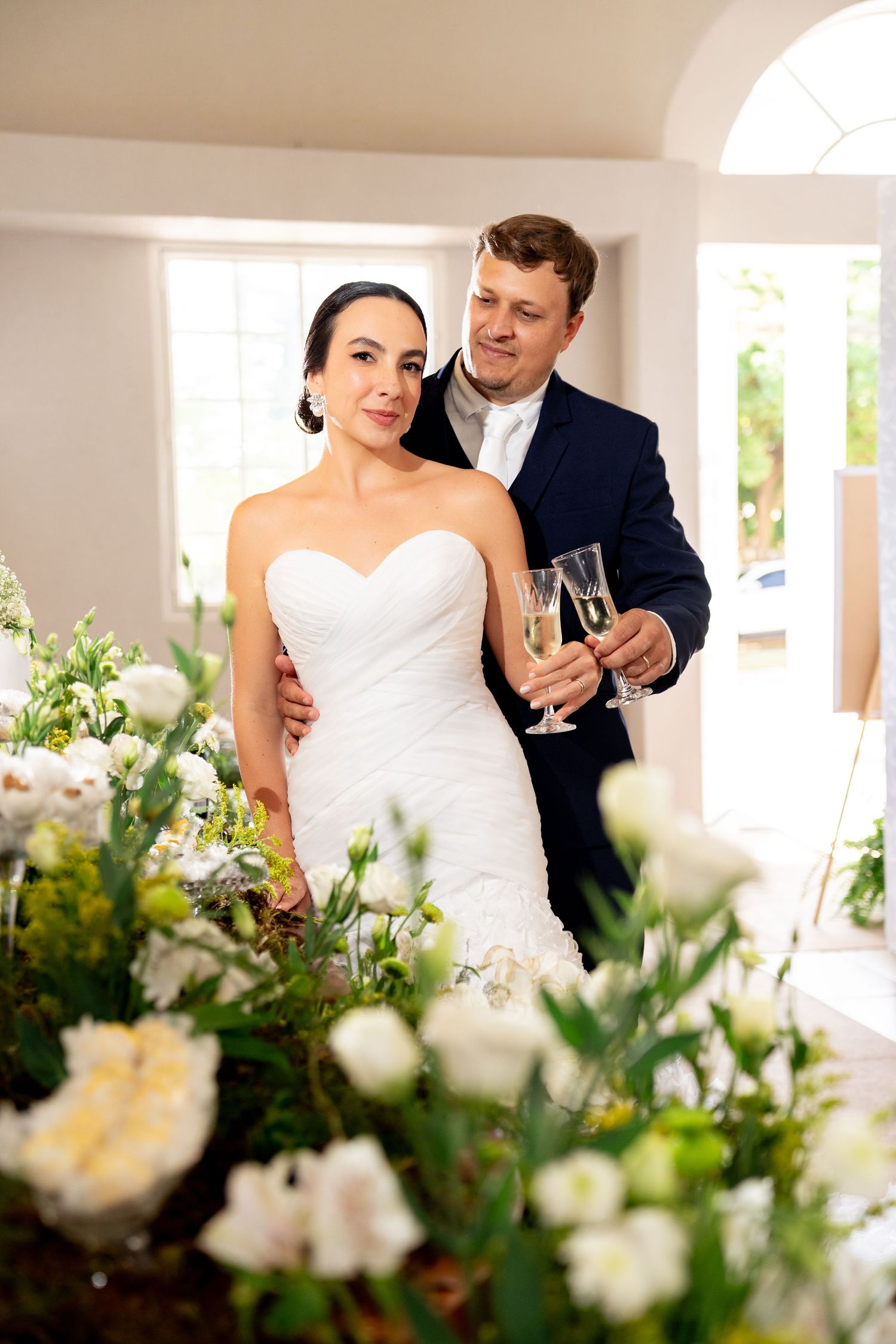 Bride and groom toasting with champagne glasses surrounded by white floral arrangements at their elegant wedding reception.