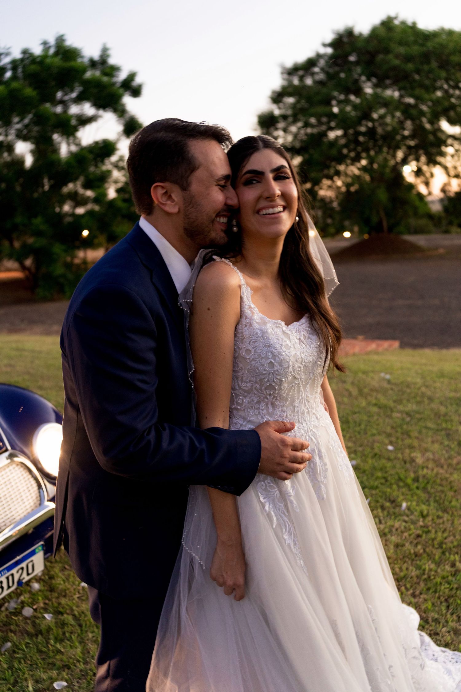 A smiling bride in a white lace gown embraces her groom in a navy suit outdoors at sunset.