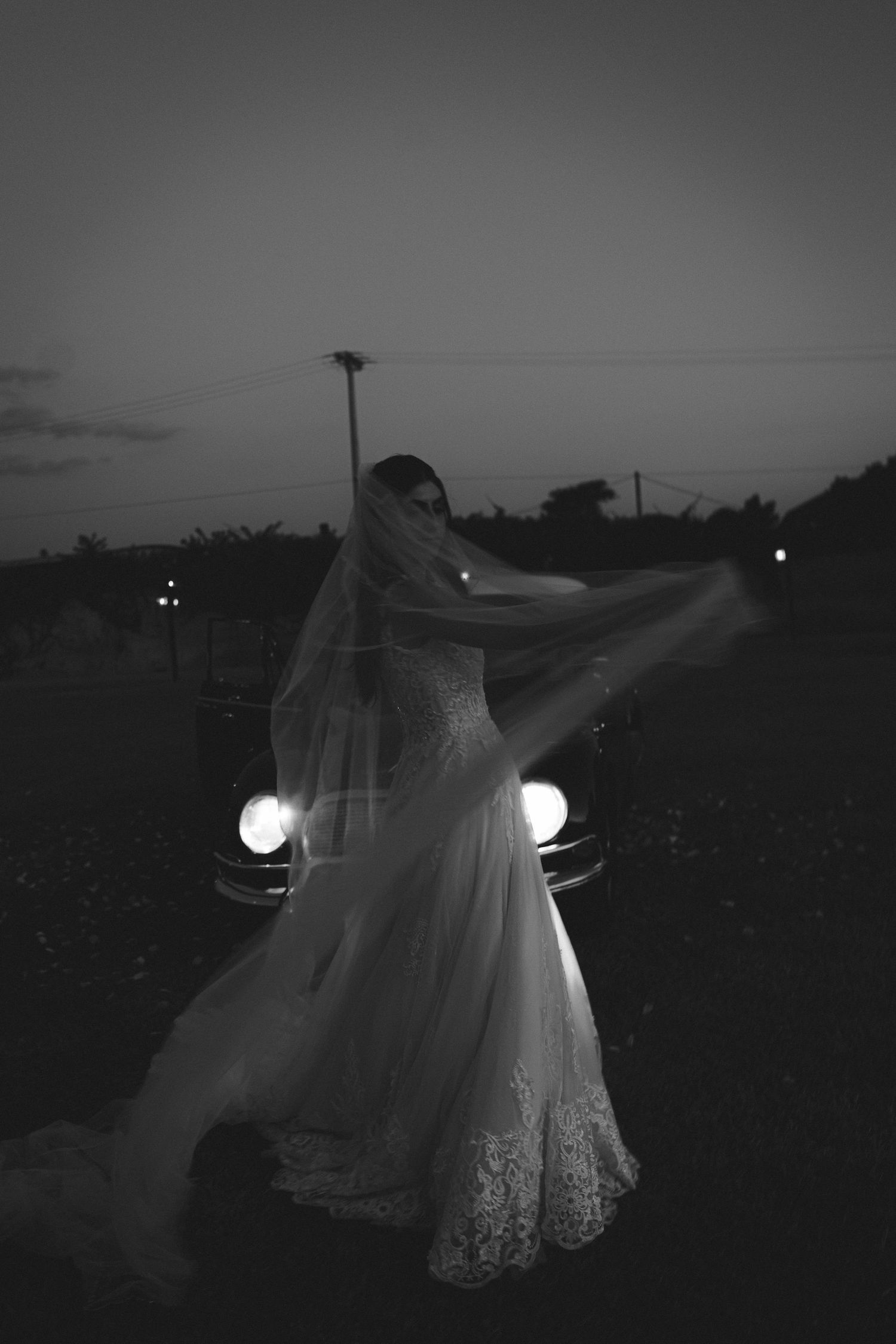 Black and white photo of a bride in a lace gown and flowing veil standing before vintage car headlights at dusk.