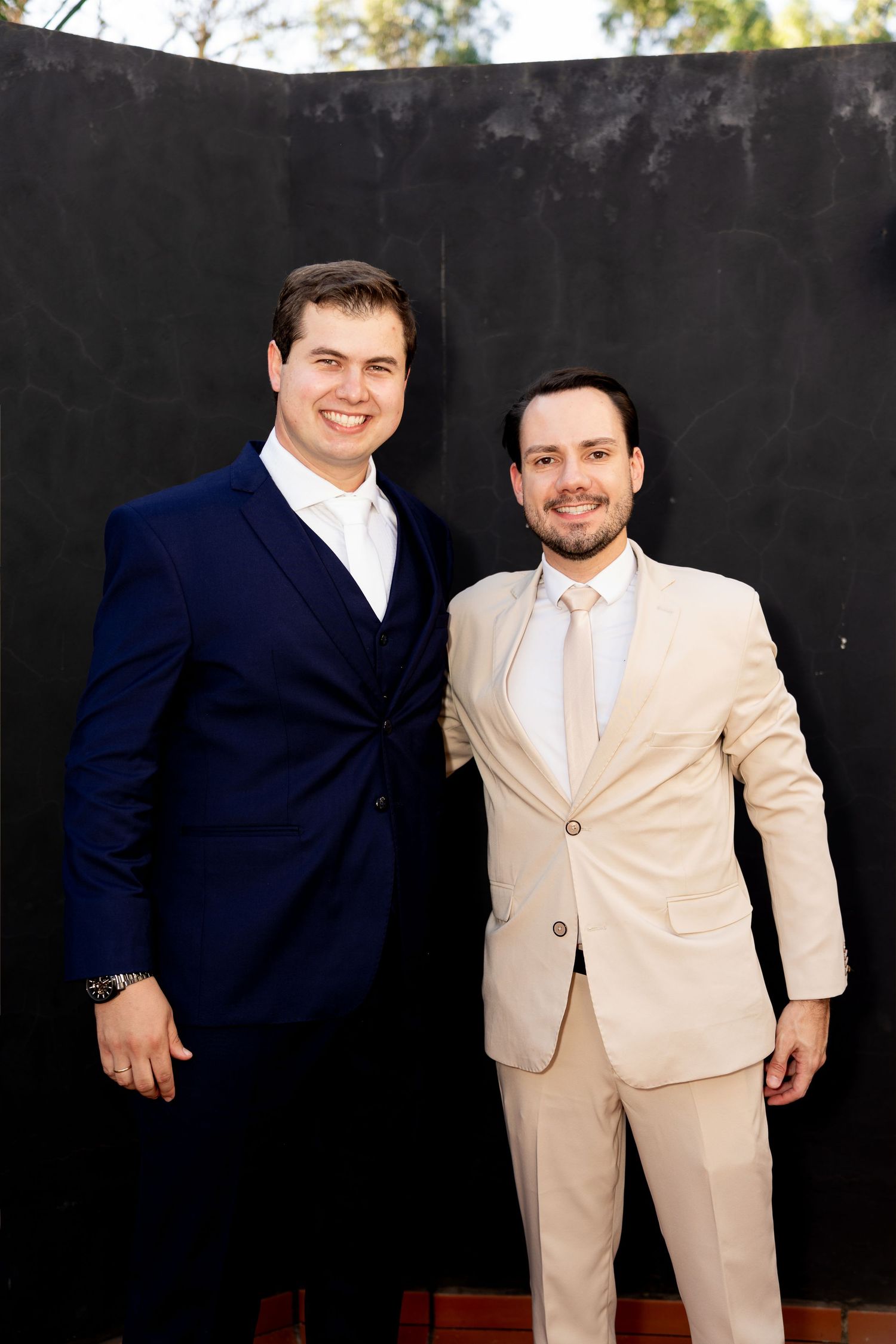 Two men in suits, one navy blue and one beige, smiling together against a dark wall backdrop.