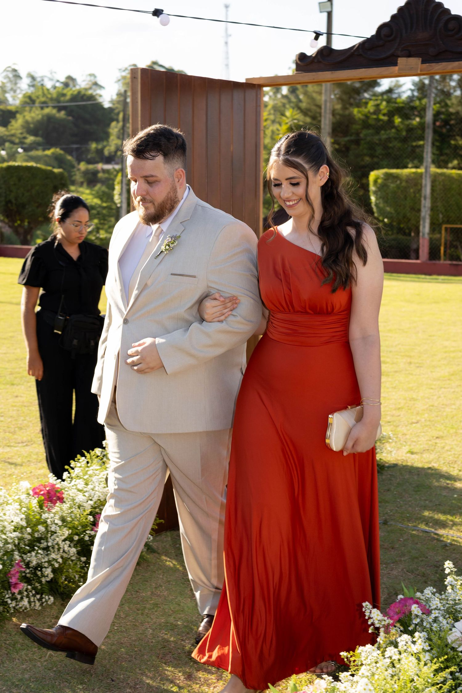 A man in a beige suit escorts a woman in a red one-shoulder gown at an outdoor wedding ceremony.