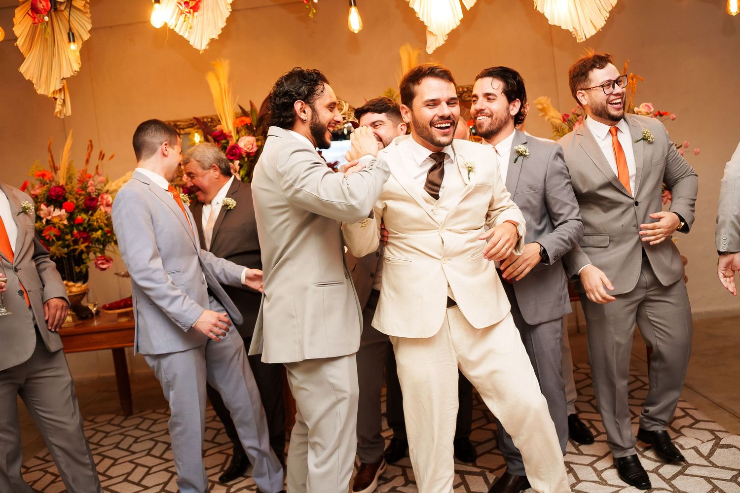 Groom in cream suit laughing with groomsmen in gray suits at a wedding reception.