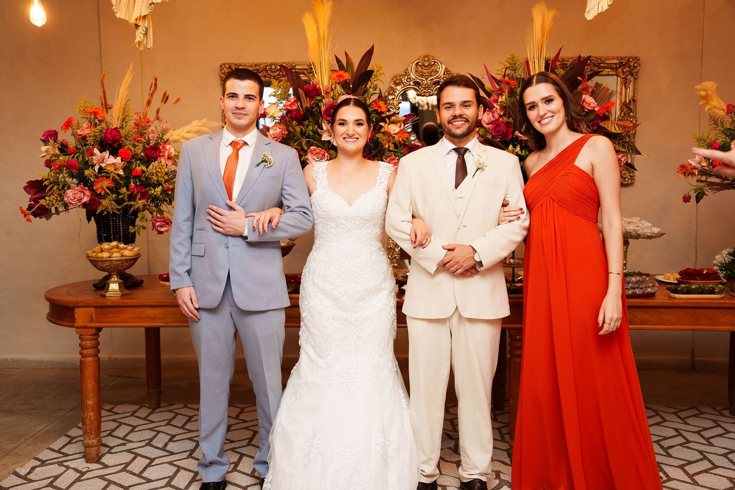 Wedding party of four posing together, bride in white gown, groom in cream suit, groomsman in gray, bridesmaid in red dress.