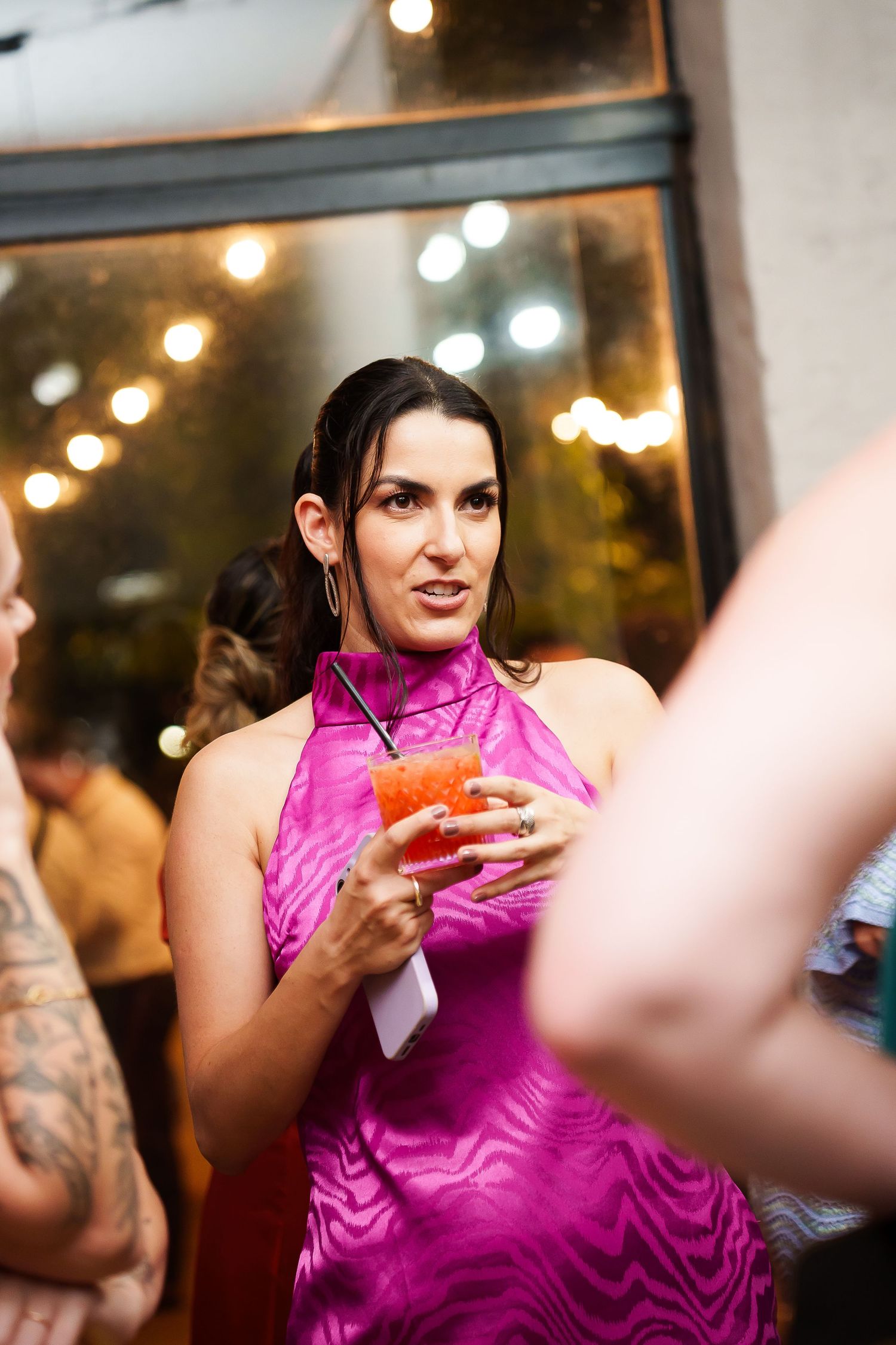 Woman in pink satin halter dress holding a cocktail at an indoor social event with string lights.