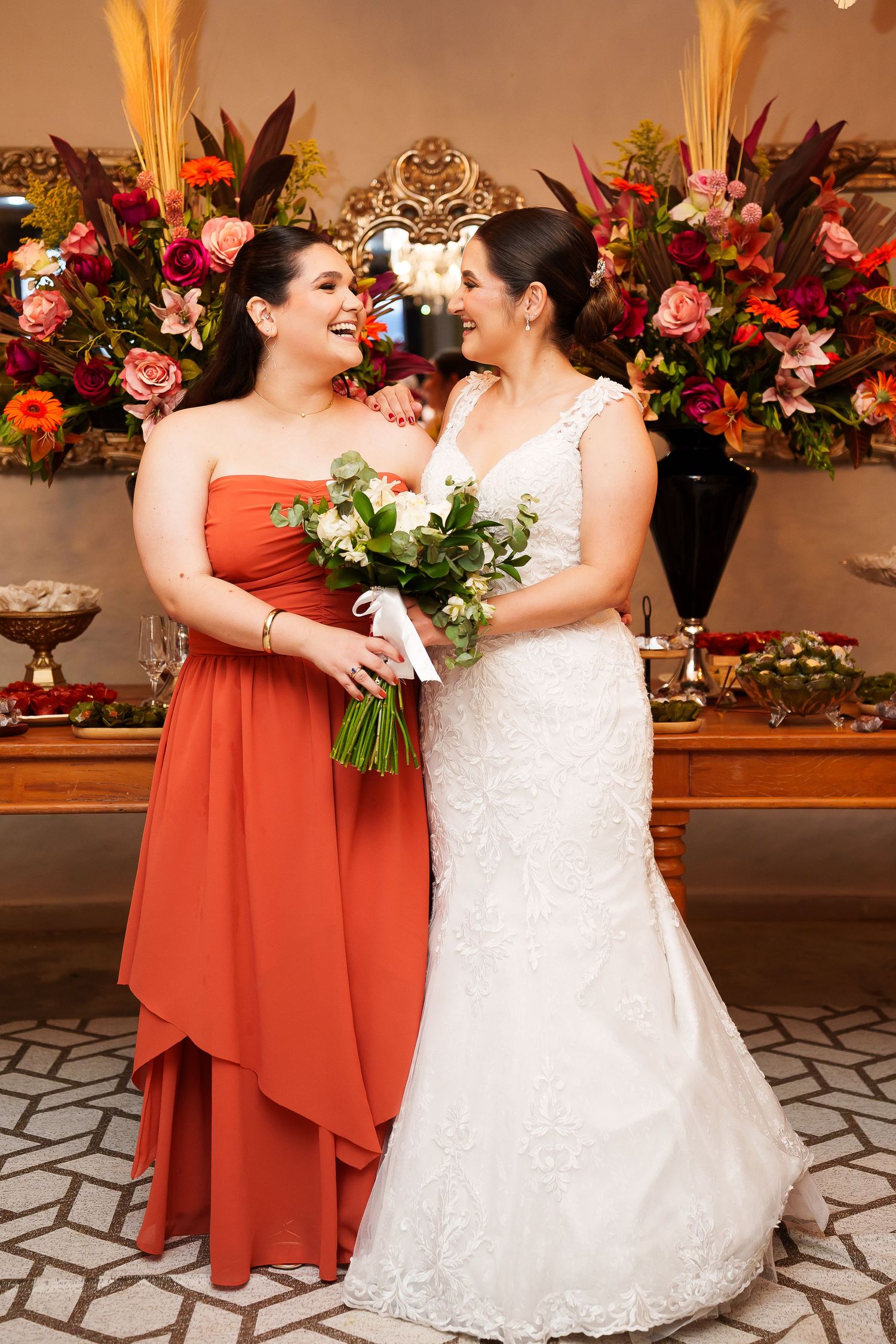 Bride in white lace gown and bridesmaid in orange strapless dress holding bouquets, smiling together at wedding reception.