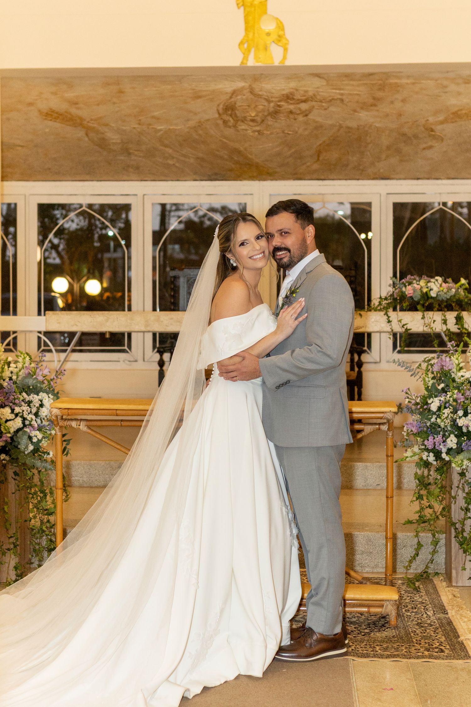 Bride in white gown and veil poses with groom in gray suit at elegant floral wedding ceremony altar.