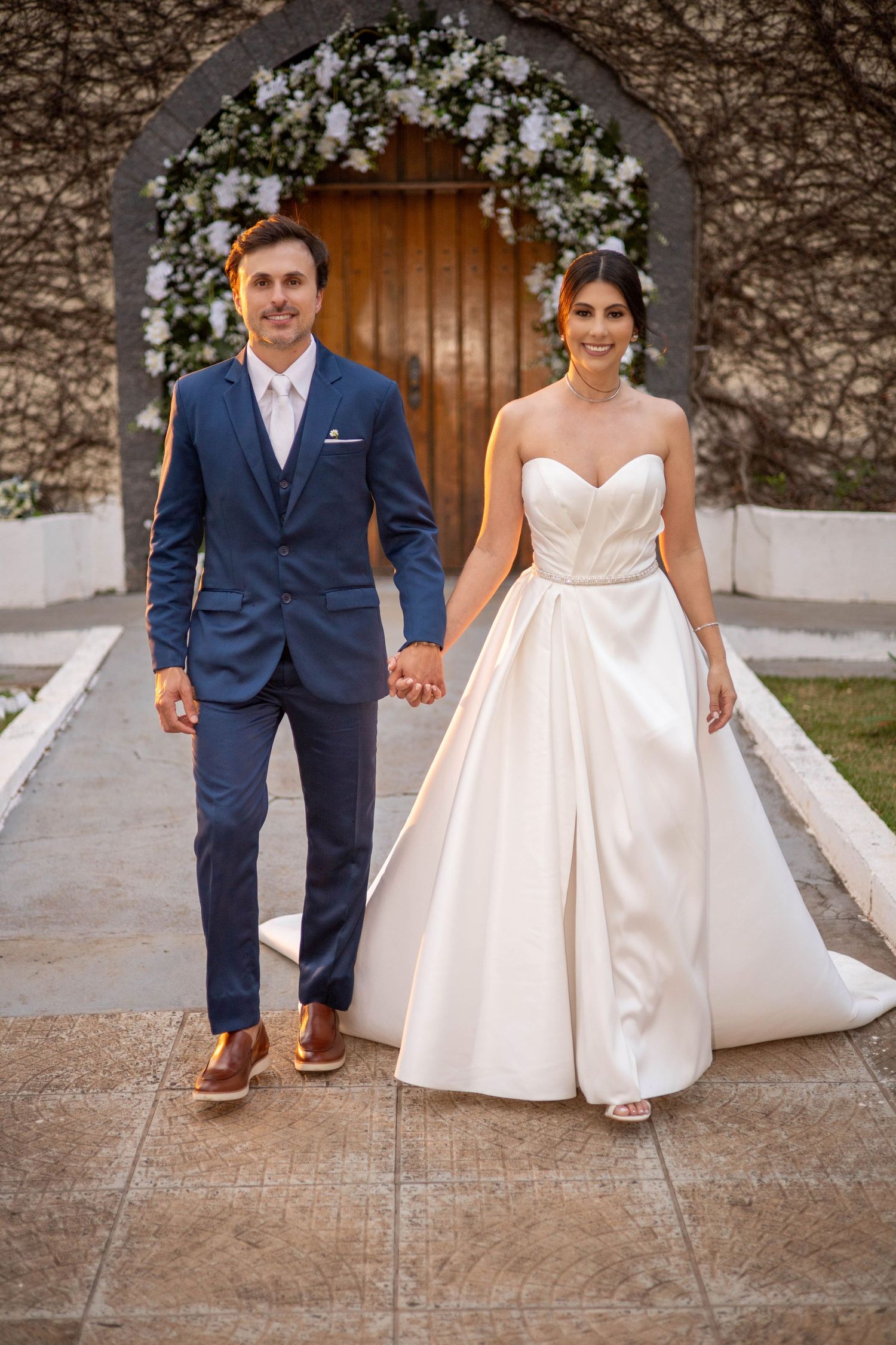 Bride in strapless satin ballgown and groom in navy suit hold hands walking outside a floral arch doorway.