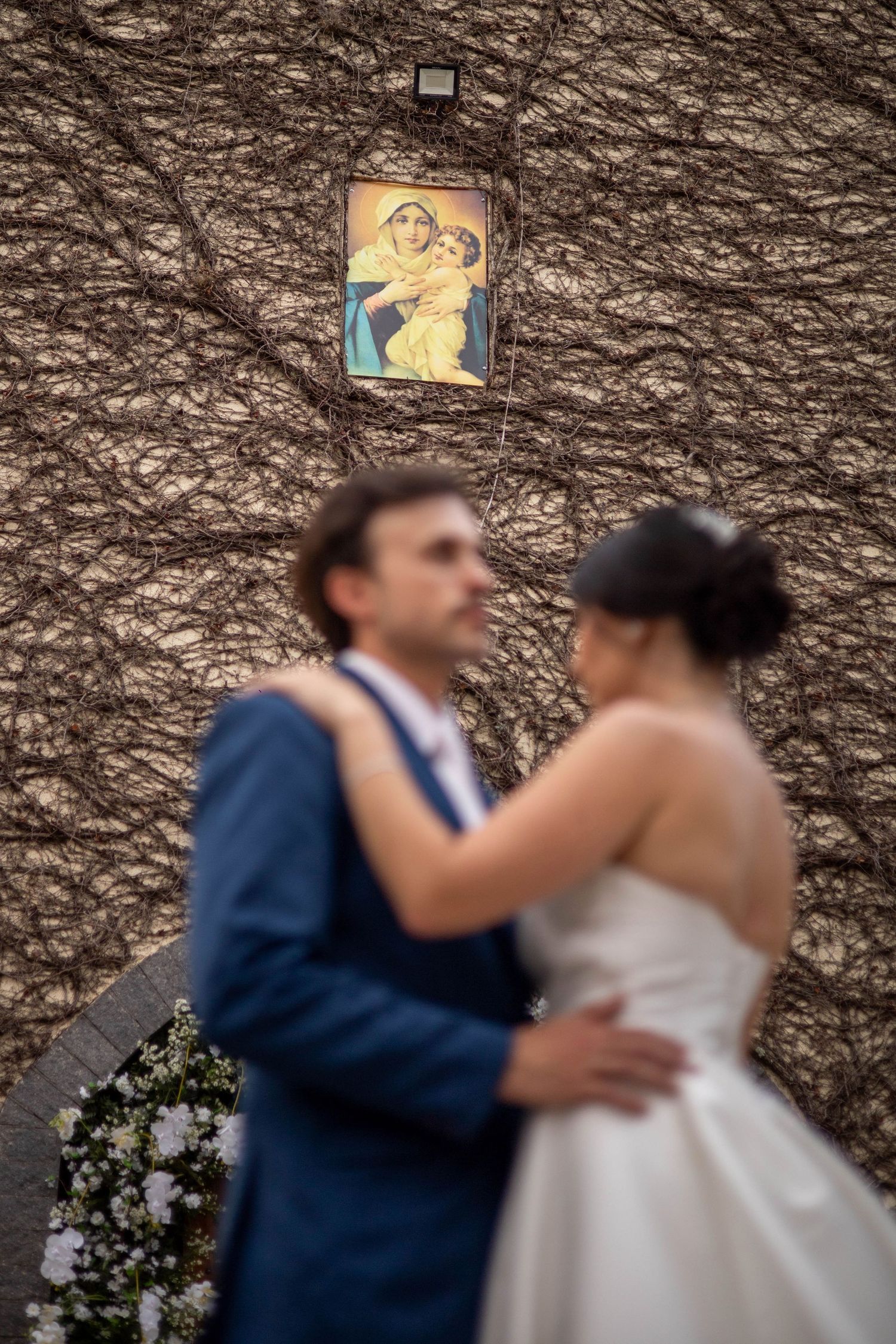 Blurred bride and groom embracing before a vine-covered wall with a religious icon of Madonna and Child above them.