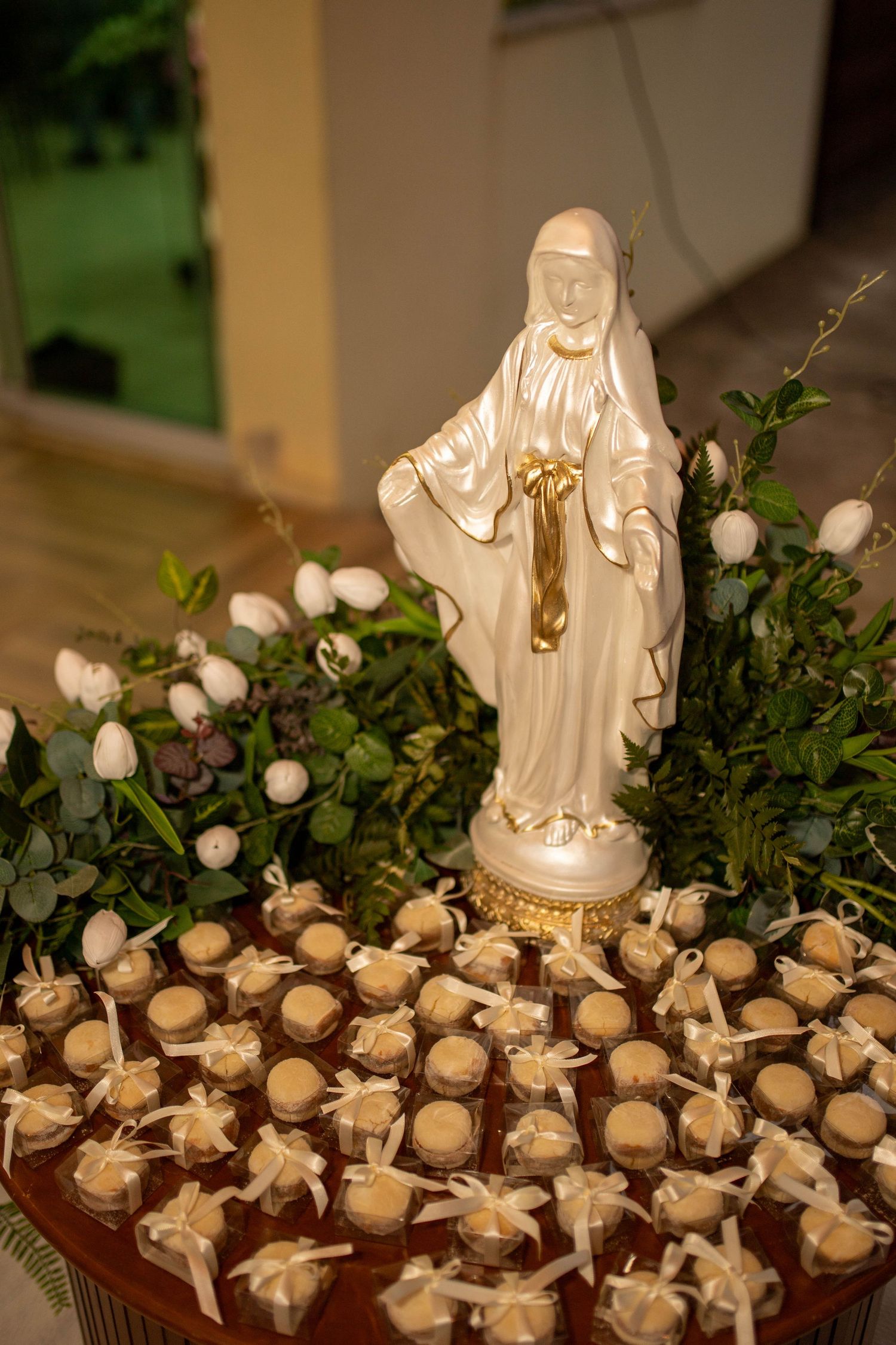 White Virgin Mary statue surrounded by floral arrangements and gold-tied favor boxes on a decorative table.