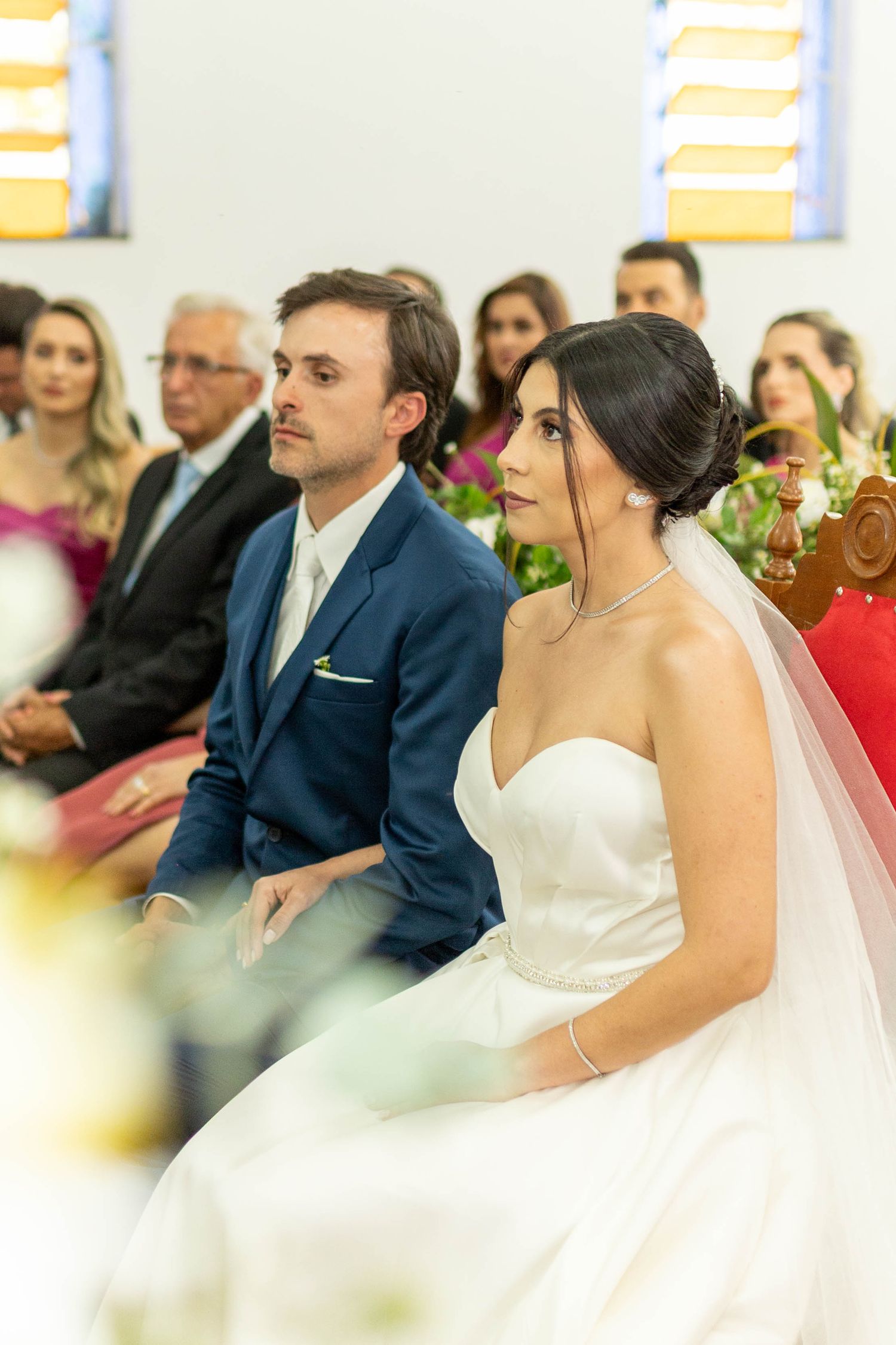 Bride in white gown and groom in blue suit seated at church wedding ceremony with guests in background.
