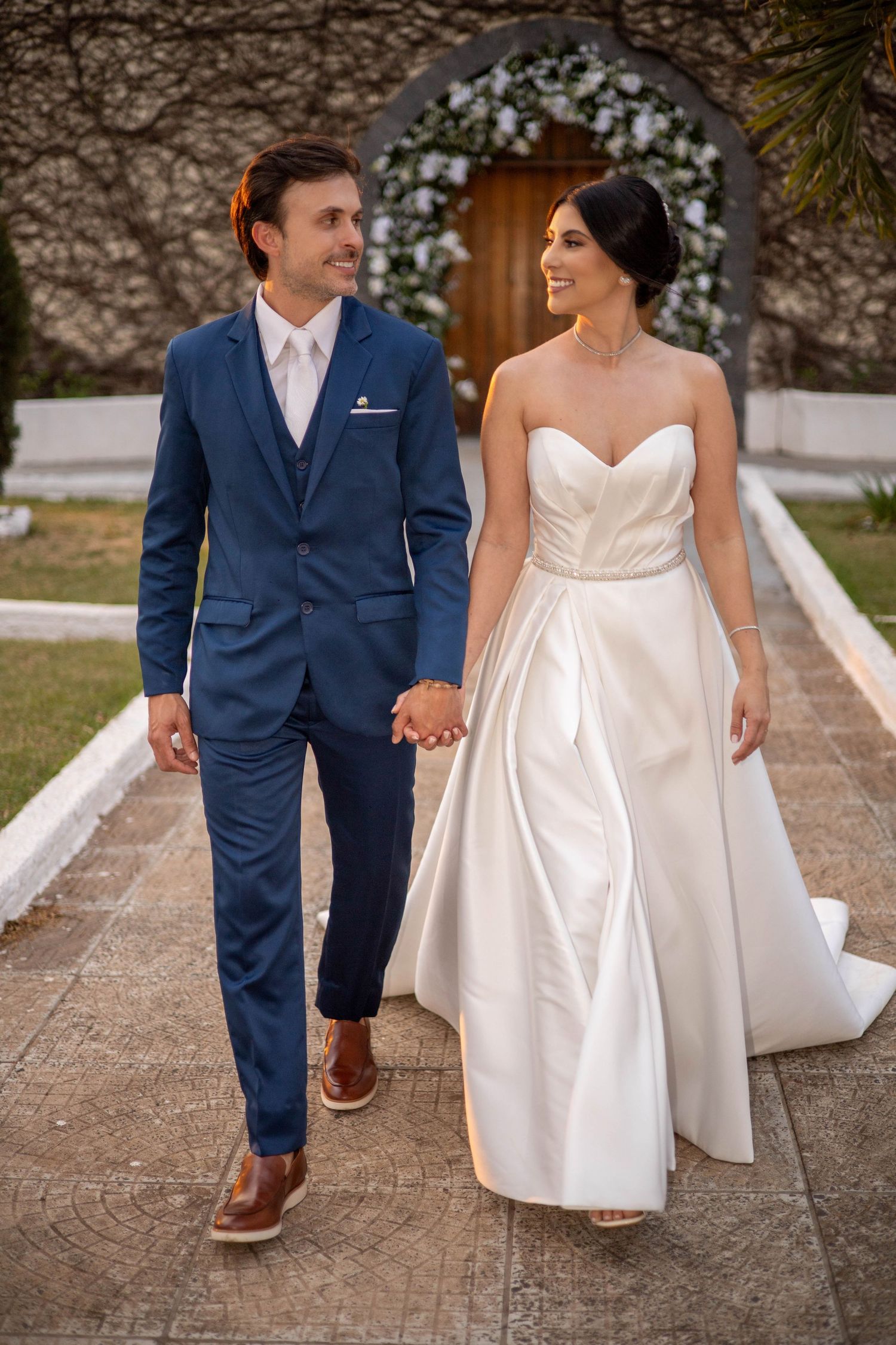 Bride in strapless white gown and groom in blue suit hold hands walking outdoors near floral arch.