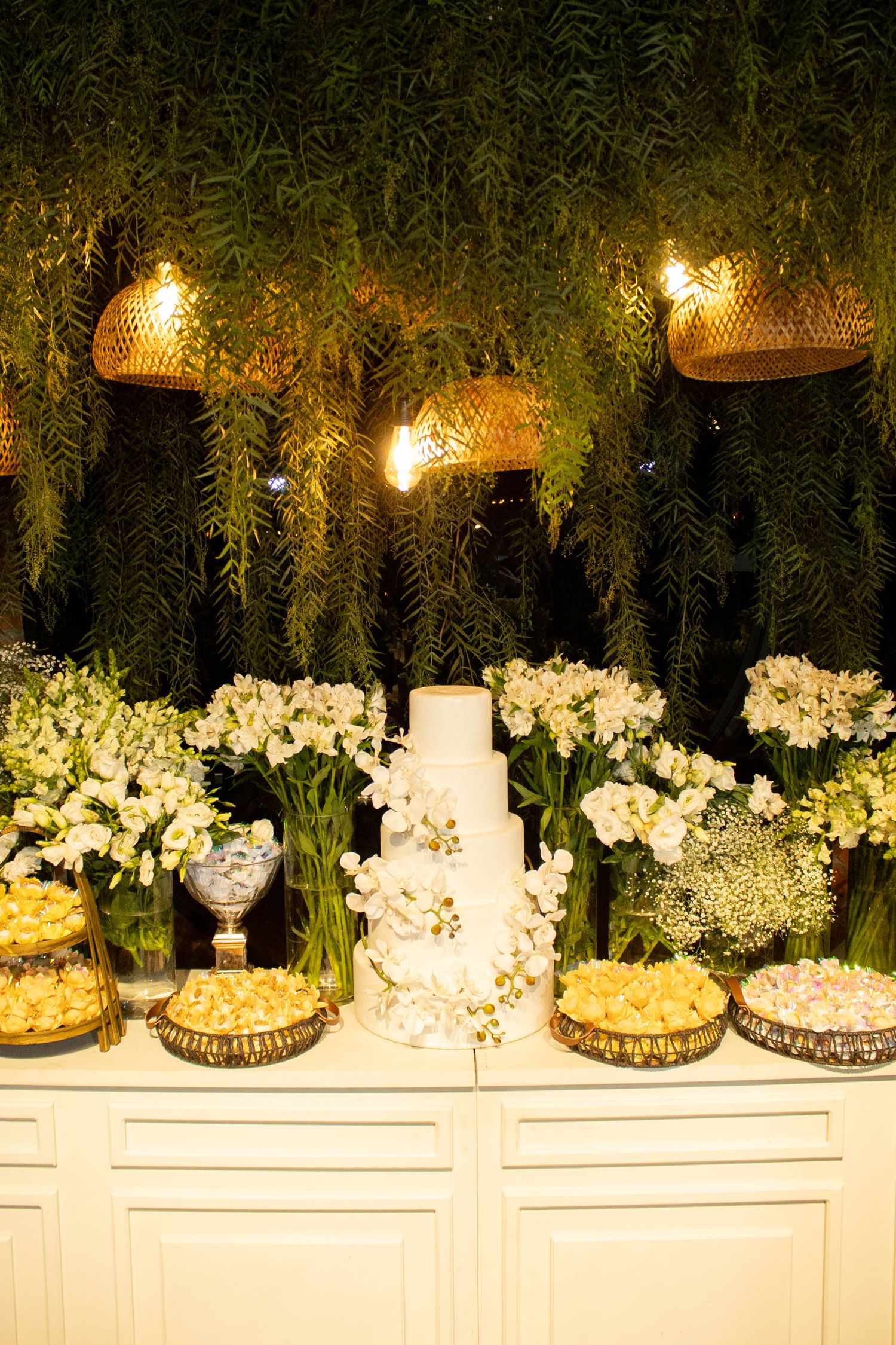 Elegant wedding dessert table with white tiered cake, fresh white flowers, and warm glowing lights against lush green backdrop.