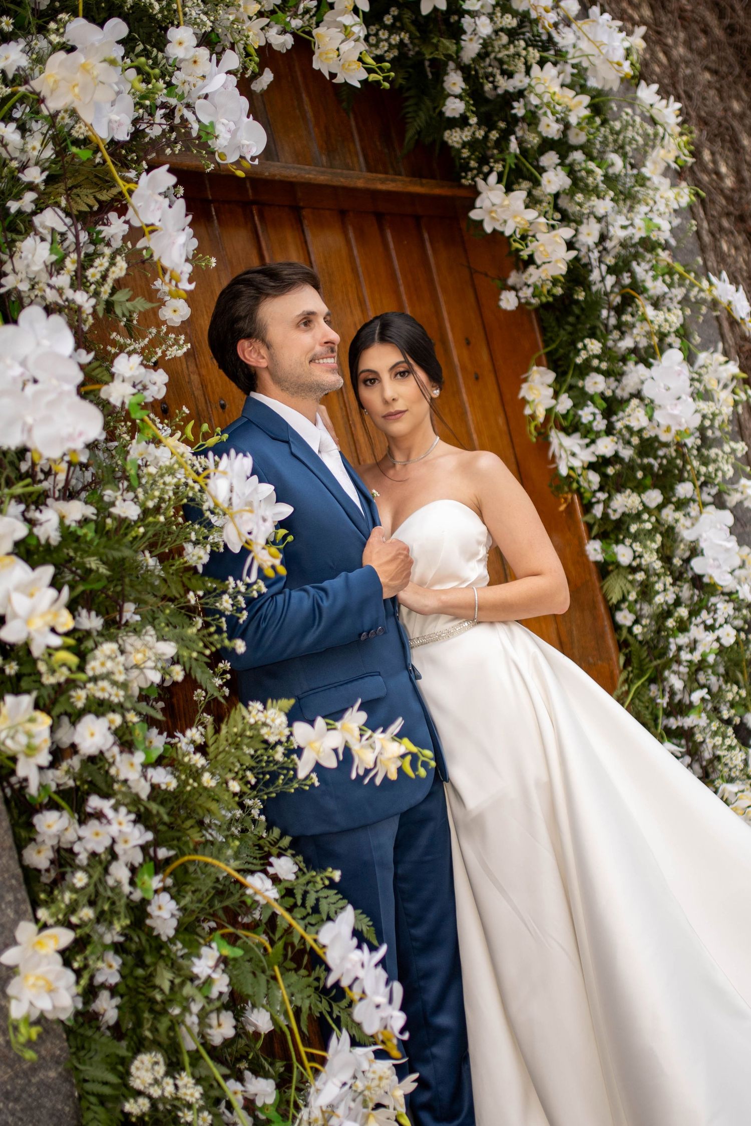 Bride and groom embracing under a stunning white floral arch with orchids at their elegant wedding ceremony.