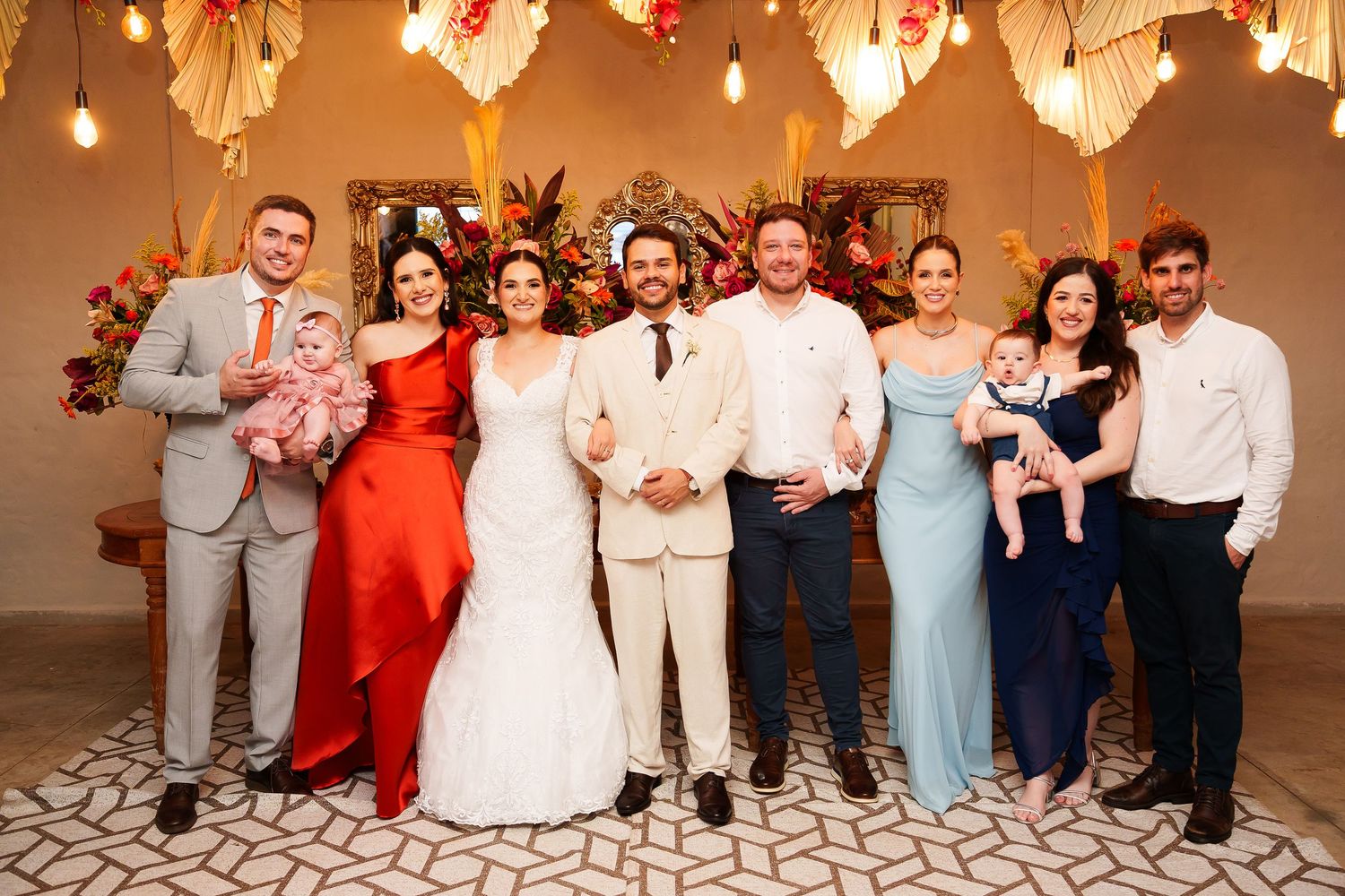 Wedding party poses together in elegant venue with floral decor, bride in white, groom in beige, guests in formal attire.