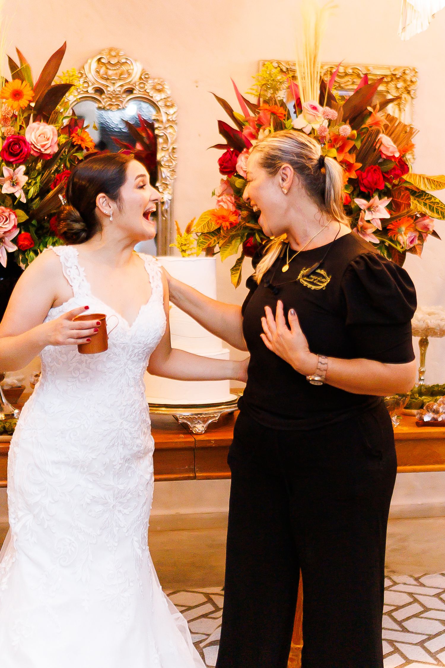 Bride in white lace gown laughing with woman in black outfit at wedding reception with floral arrangements.