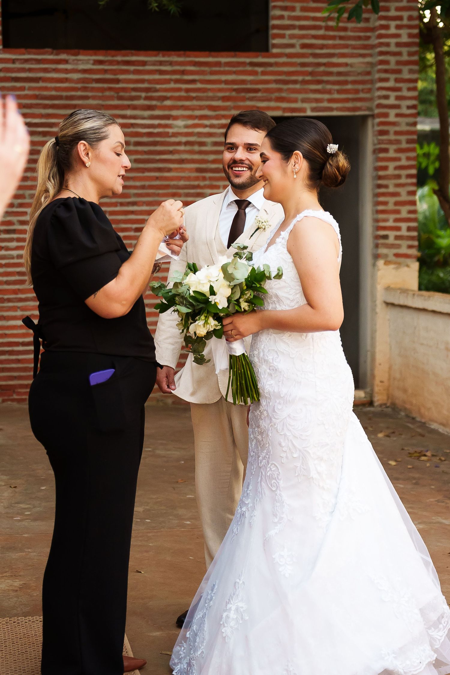 Bride in white lace gown holds bouquet while groom in cream suit smiles as woman in black adjusts details outdoors.