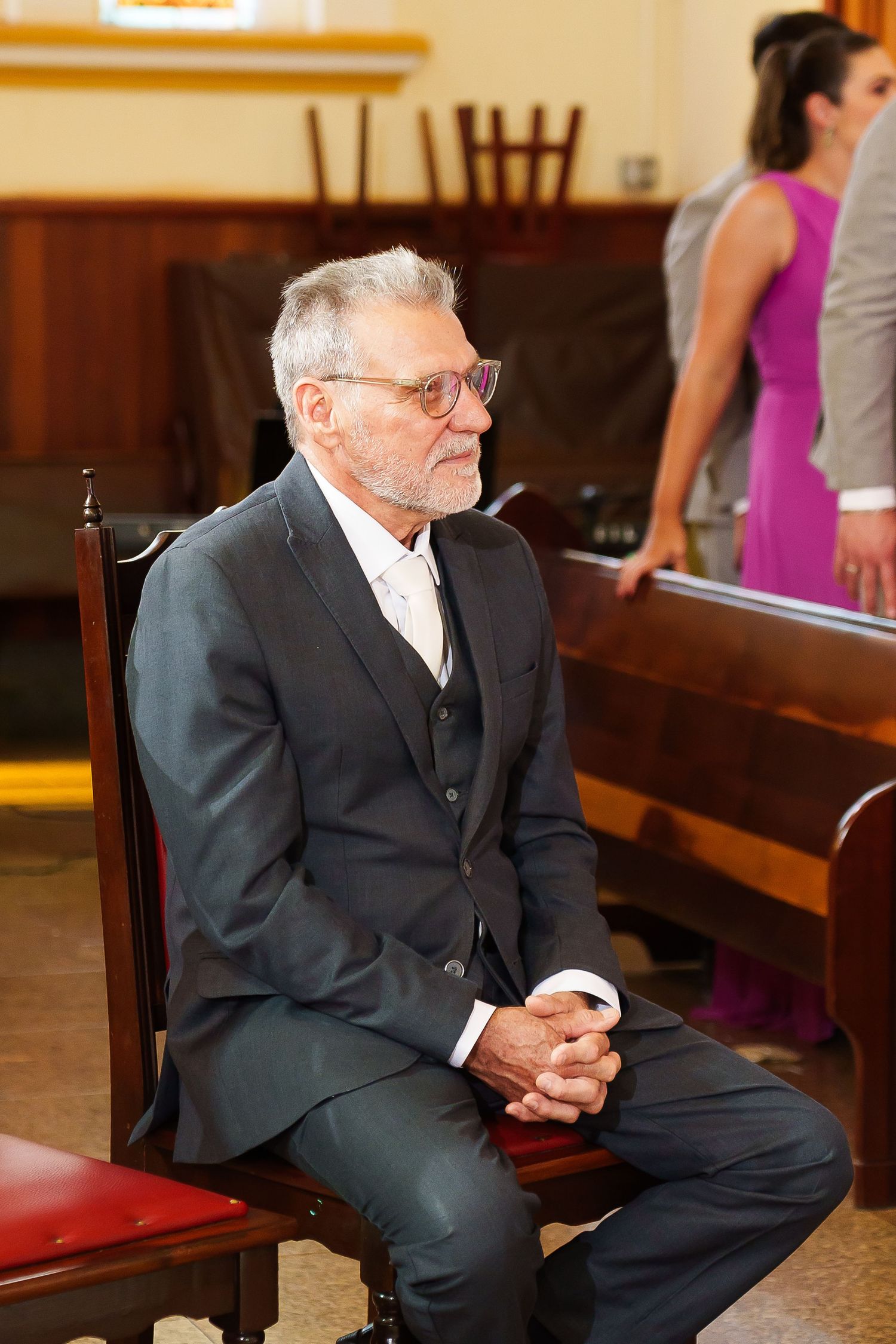 Elderly man in gray suit sitting in church pew with hands clasped, wearing glasses at a formal event.