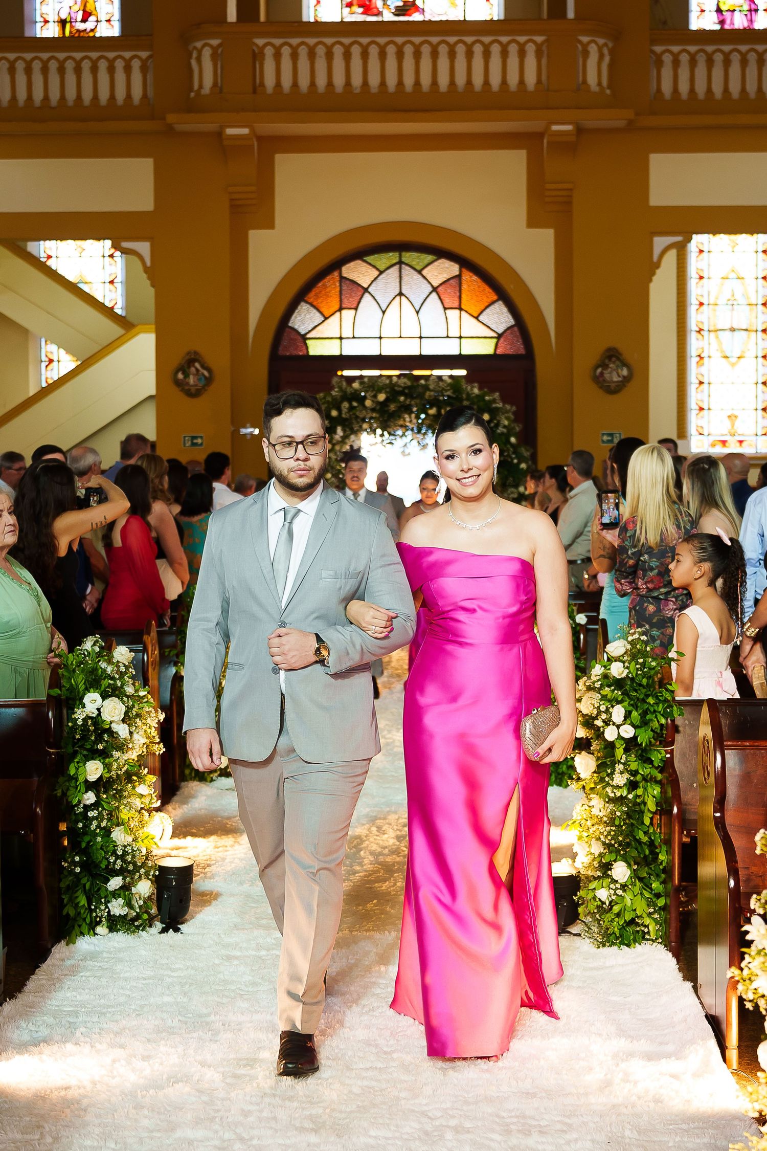 A couple walks down a church aisle, man in gray suit, woman in pink strapless gown, at a wedding ceremony.