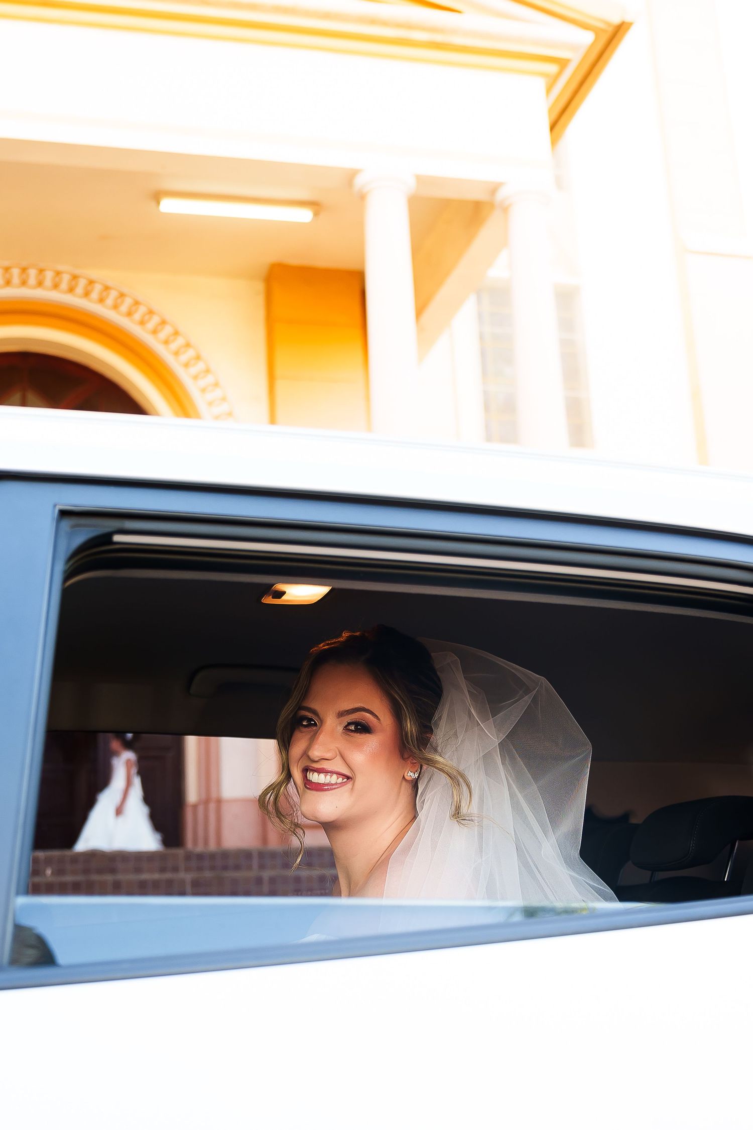 Smiling bride in veil looking out of white limousine window at wedding venue with elegant columns.