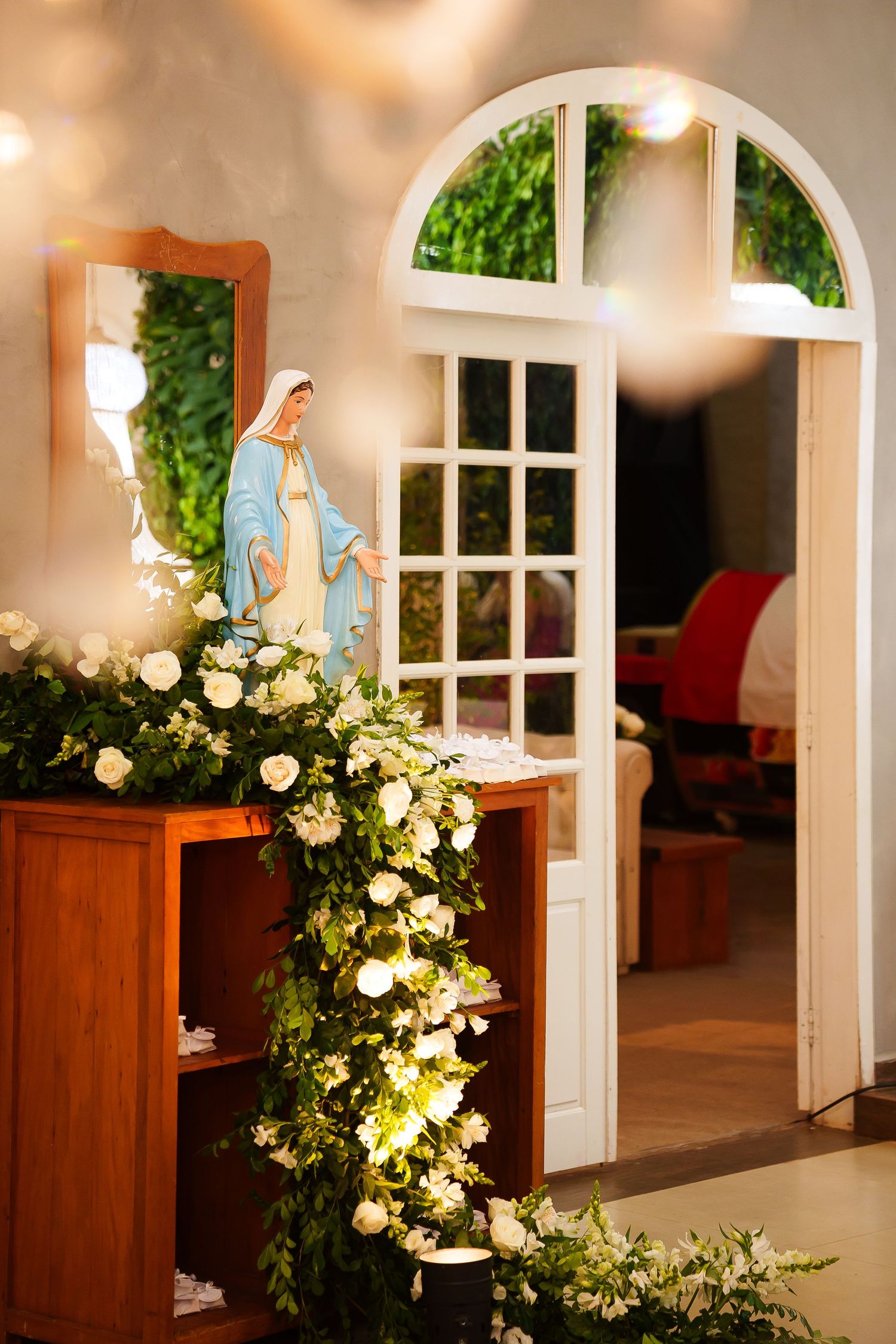 A Virgin Mary statue adorned with white roses and greenery near an arched white door in a chapel.