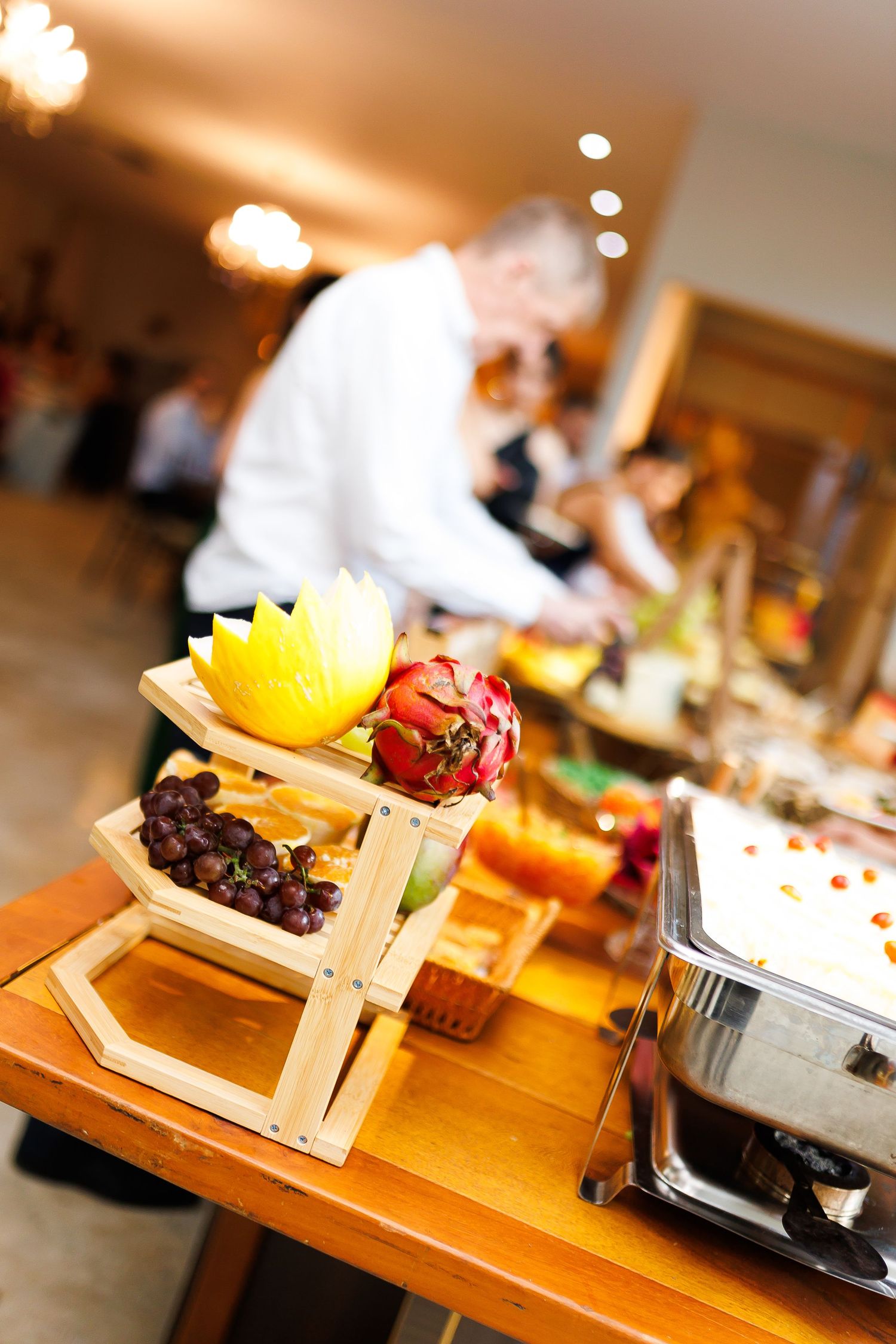 Elegant buffet display with tiered wooden stand holding fresh fruits including grapes and dragon fruit at a catered event.