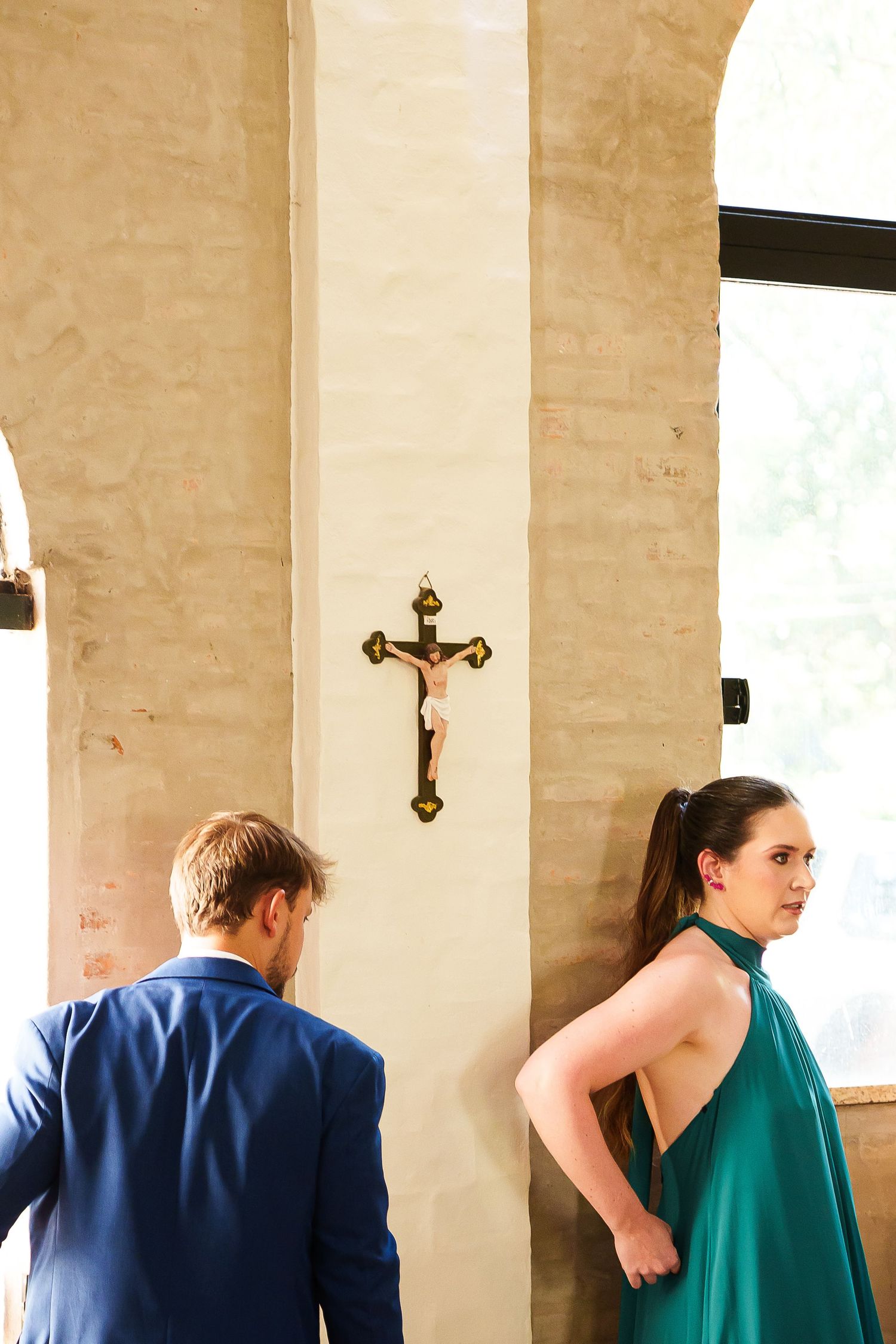 Elegant couple in formal attire standing near a crucifix on a stone wall in a sunlit church.