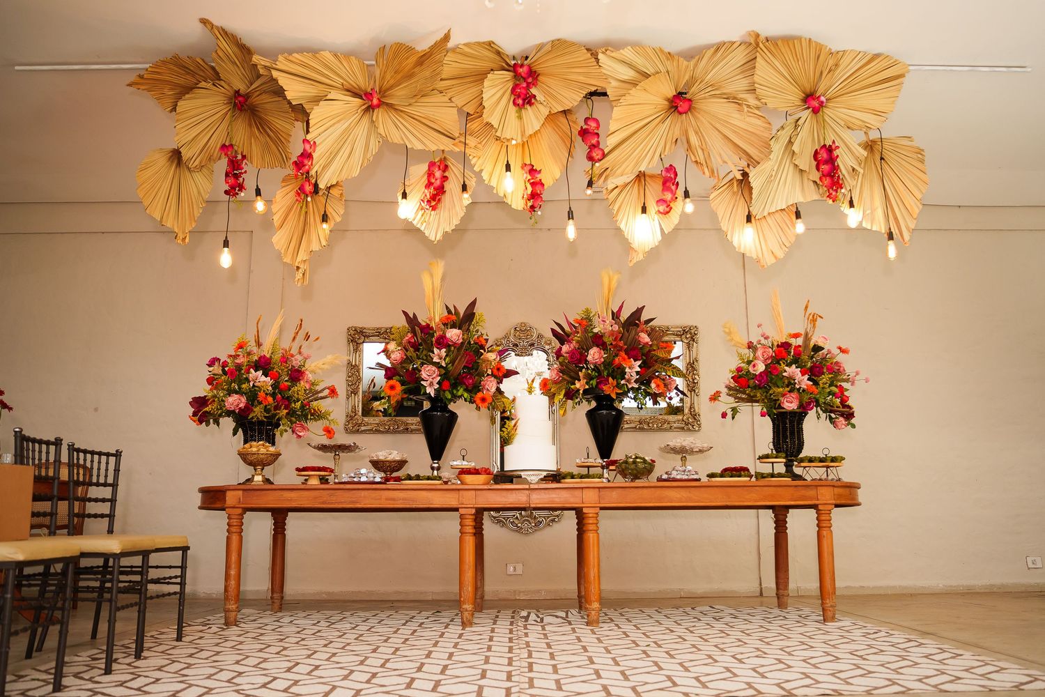 Elegant wedding reception table with tropical leaf canopy, hanging lights, and vibrant floral arrangements in warm tones.