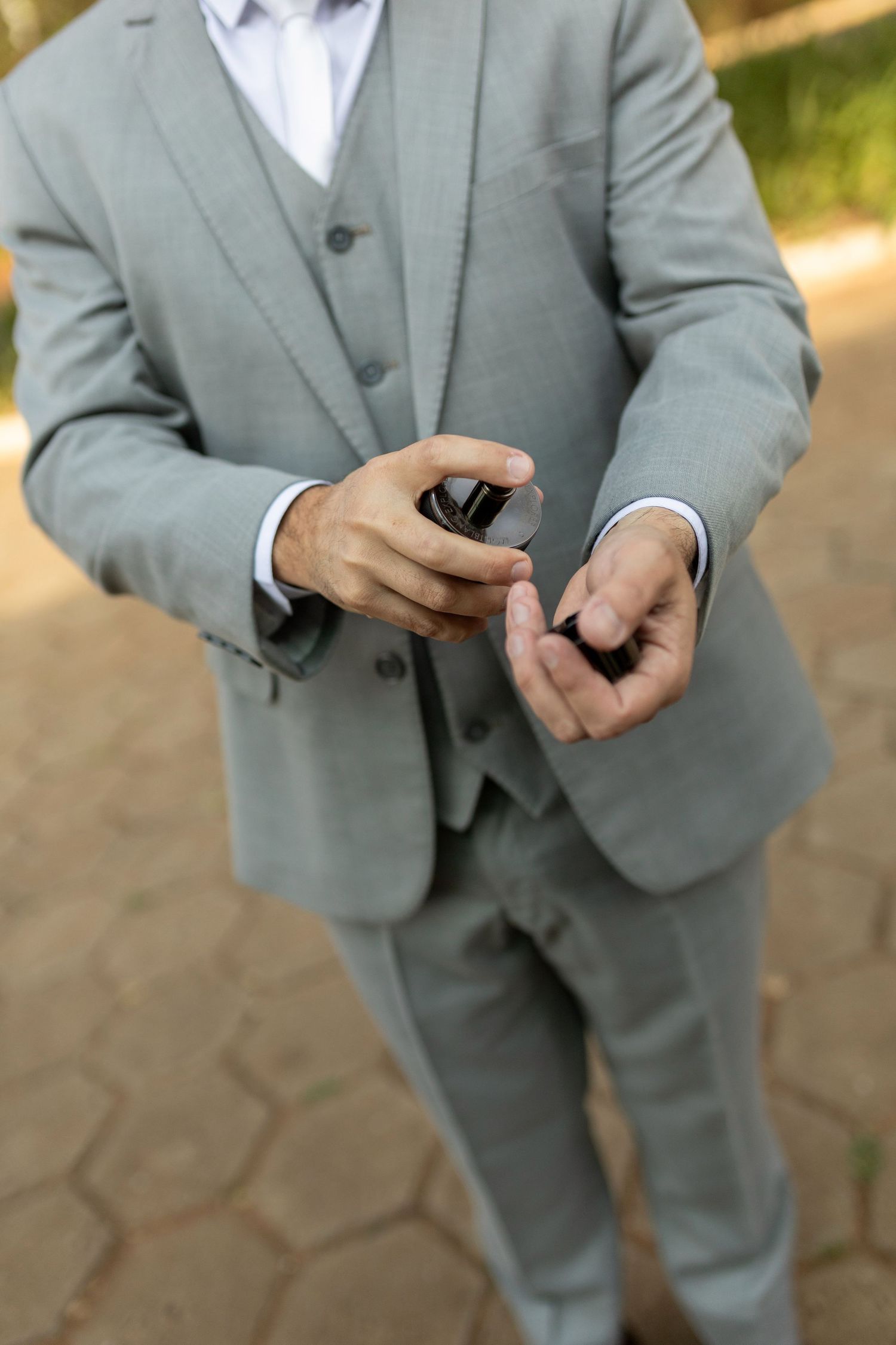 A groom in a light gray three-piece suit holds a small ring box outdoors on a paved path.