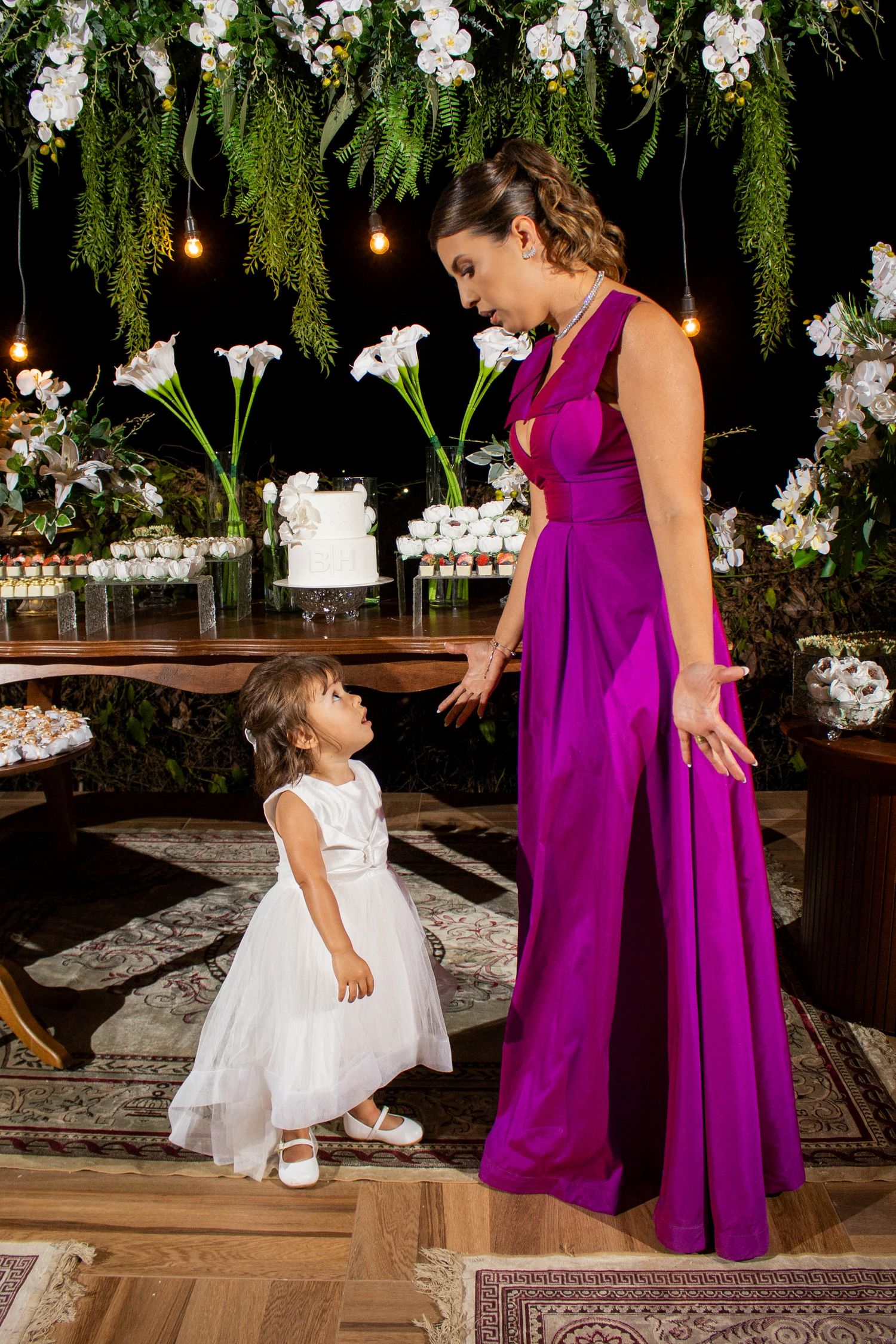 Woman in purple gown smiles at toddler in white dress at elegant floral wedding reception.
