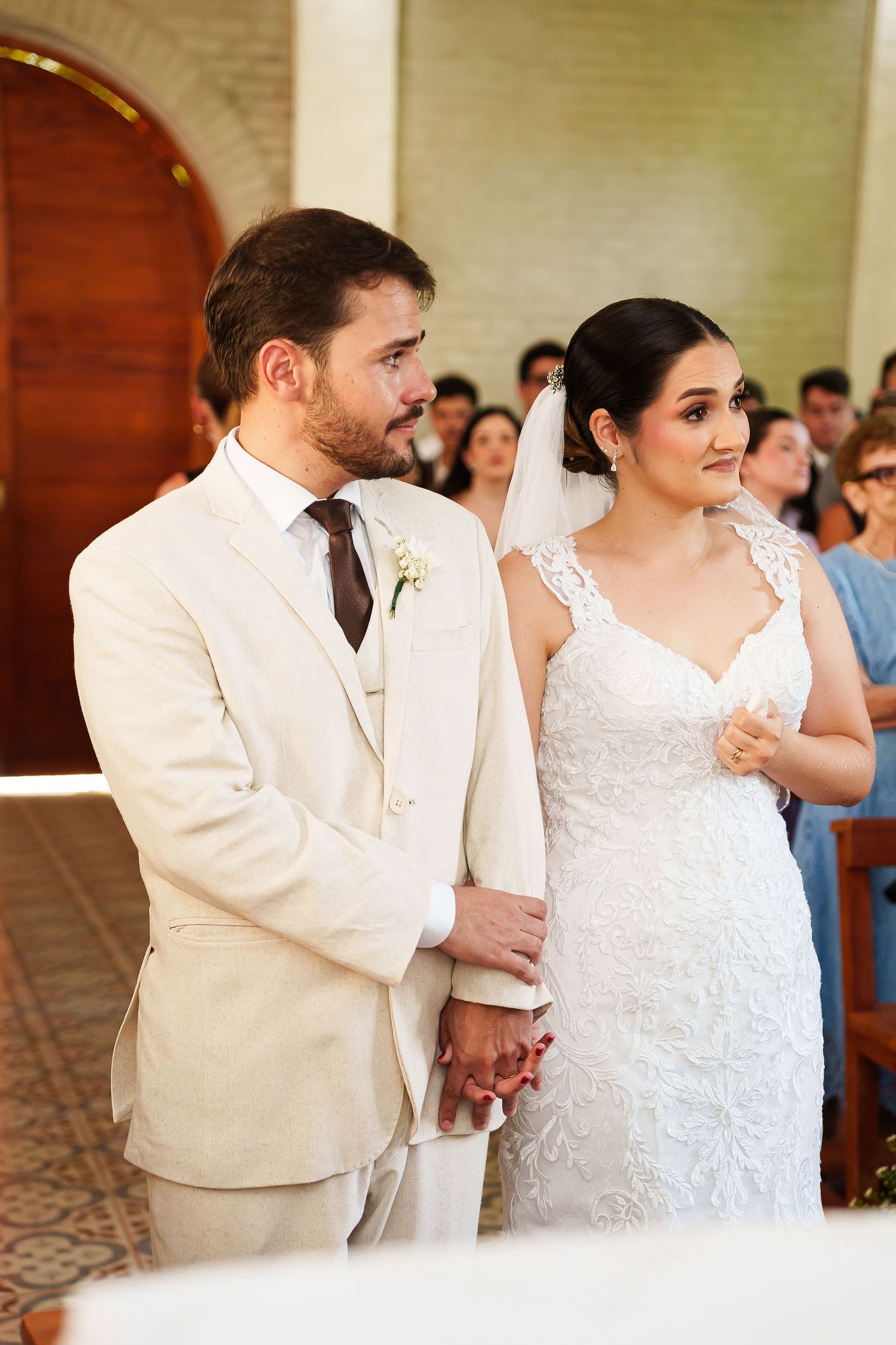 Bride in white lace gown and groom in cream suit hold hands at church wedding ceremony altar.