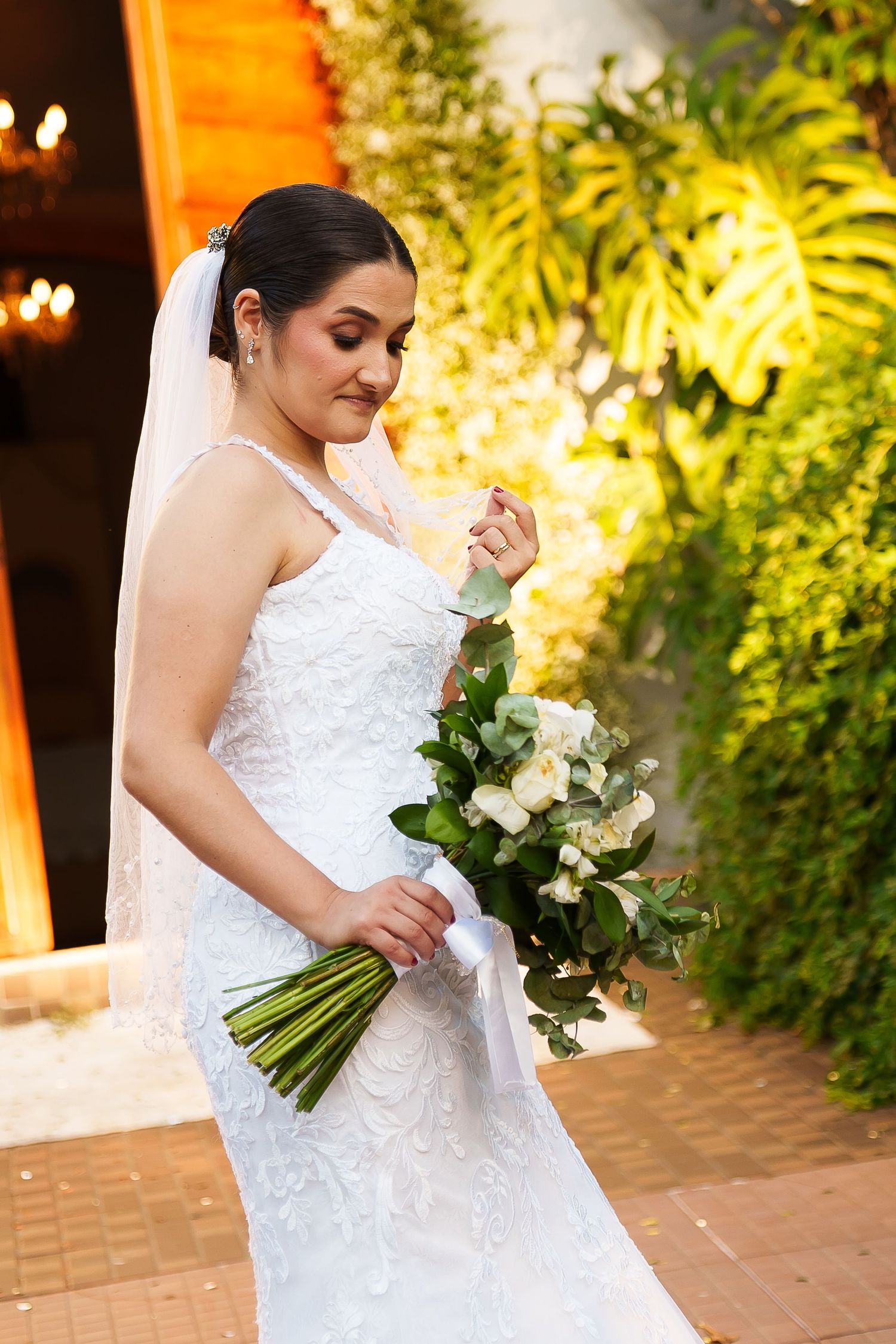 Elegant bride in white lace gown and veil holds bouquet of white roses outdoors at evening wedding venue.