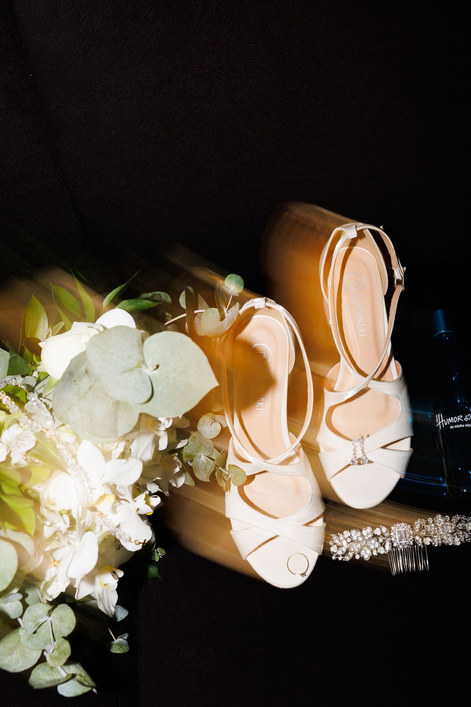Elegant bridal flat shoes with ankle straps beside a white floral bouquet and crystal jewelry on a dark background.