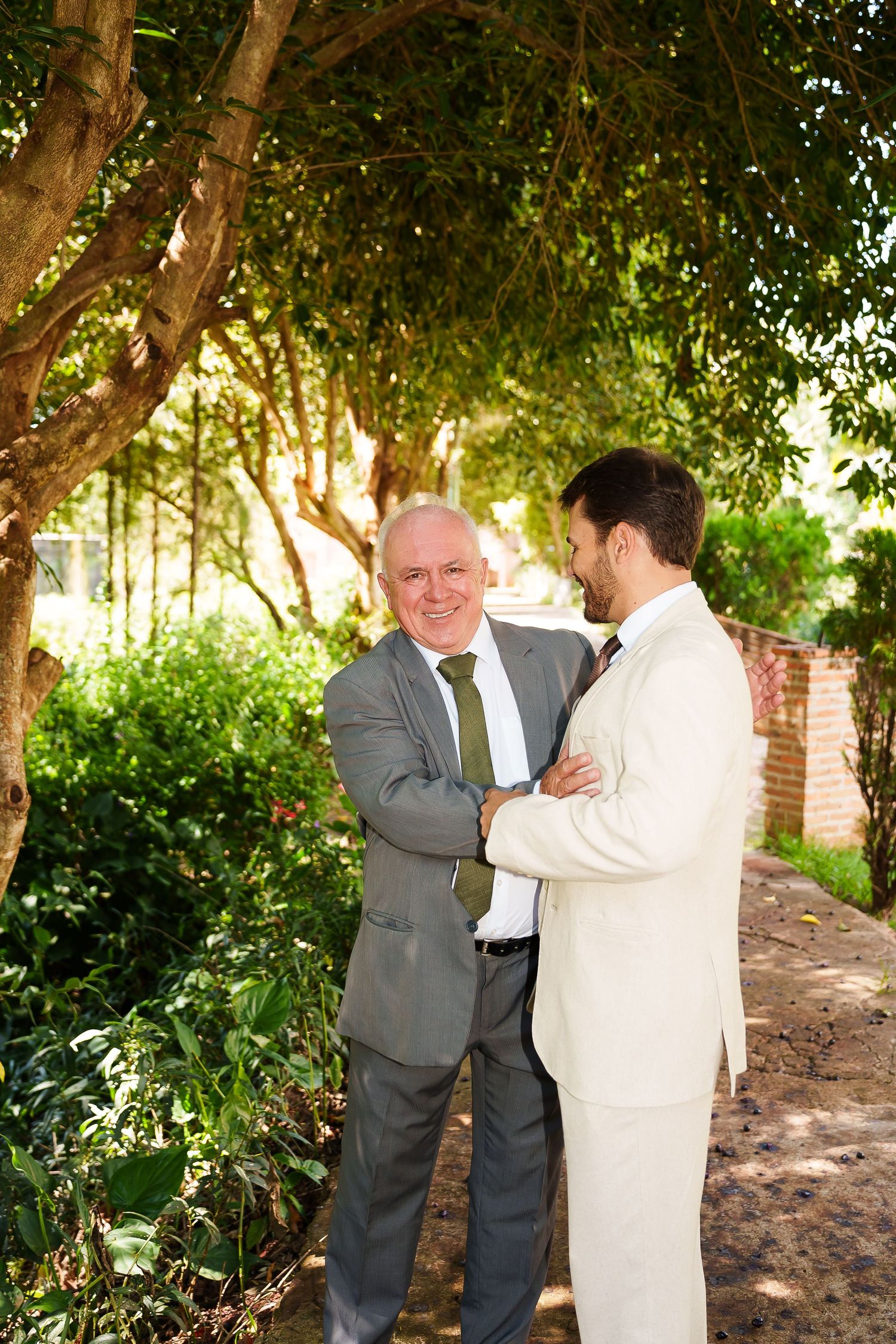 Two men in suits share a moment outdoors, with lush green trees and a brick wall in the background.