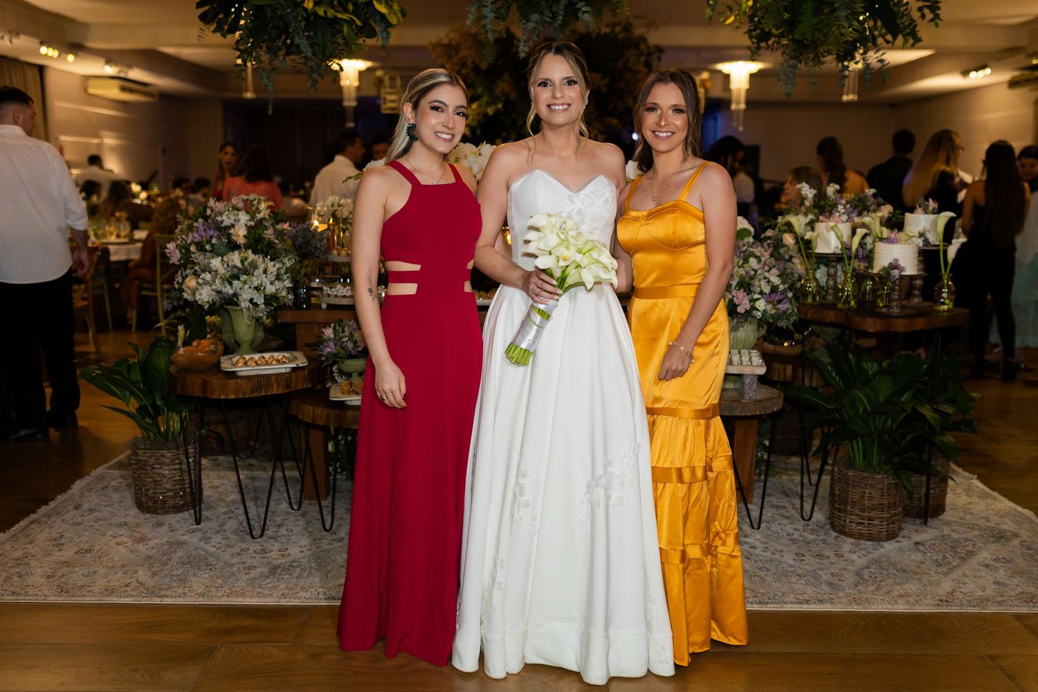 Bride in white gown poses with two bridesmaids in red and gold dresses at elegant wedding reception.