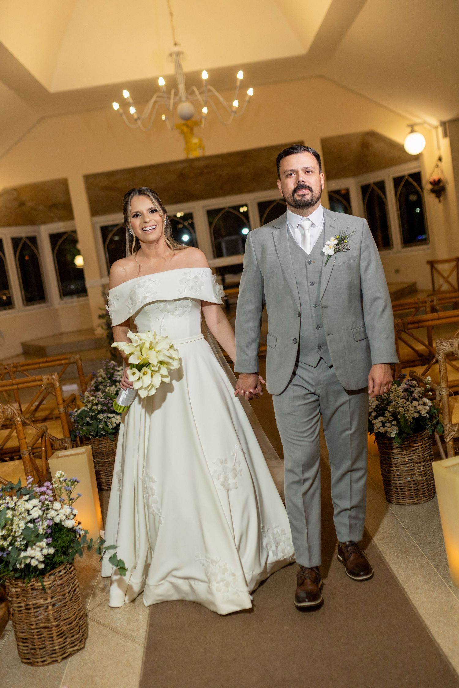 Bride in white off-shoulder gown and groom in gray suit walk hand-in-hand down church aisle after wedding ceremony.