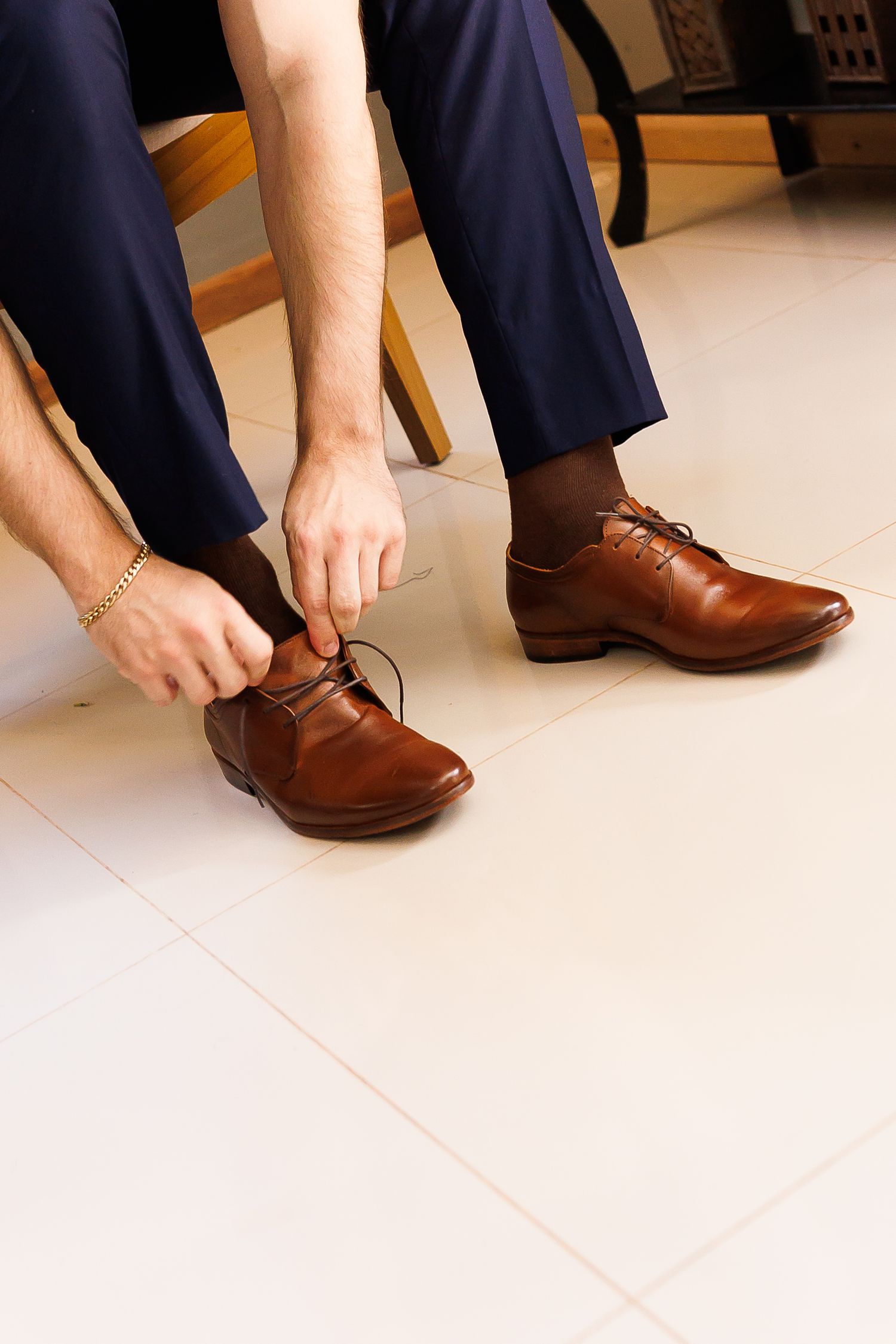 Man in navy trousers tying brown leather Oxford dress shoes while seated on wooden chair.