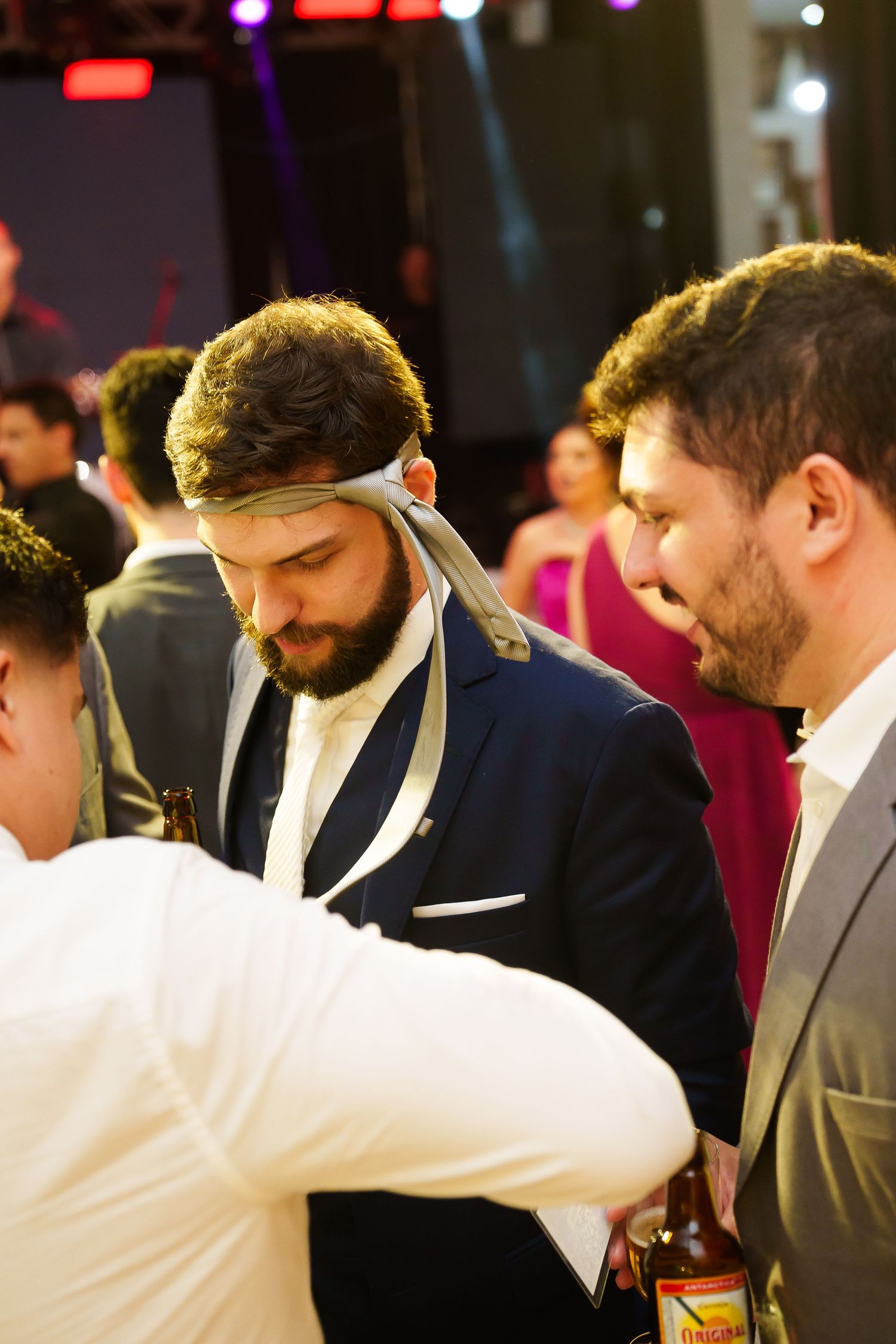 Men in formal attire at an evening event, one wearing a tie as headband holding a beer bottle.